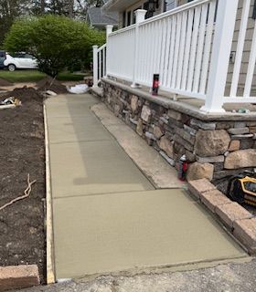Concrete sidewalk ramp beside a stone porch with white railing and landscaped yard