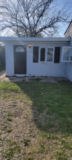 Small white house exterior with a gray door, front windows, and a patchy lawn in daylight