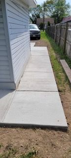 Narrow concrete side walkway beside a house, bordered by a wooden fence and grass, leading to a car in the driveway