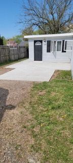 Small white backyard shed with a black door and ramp, beside a fenced grassy yard under a blue sky