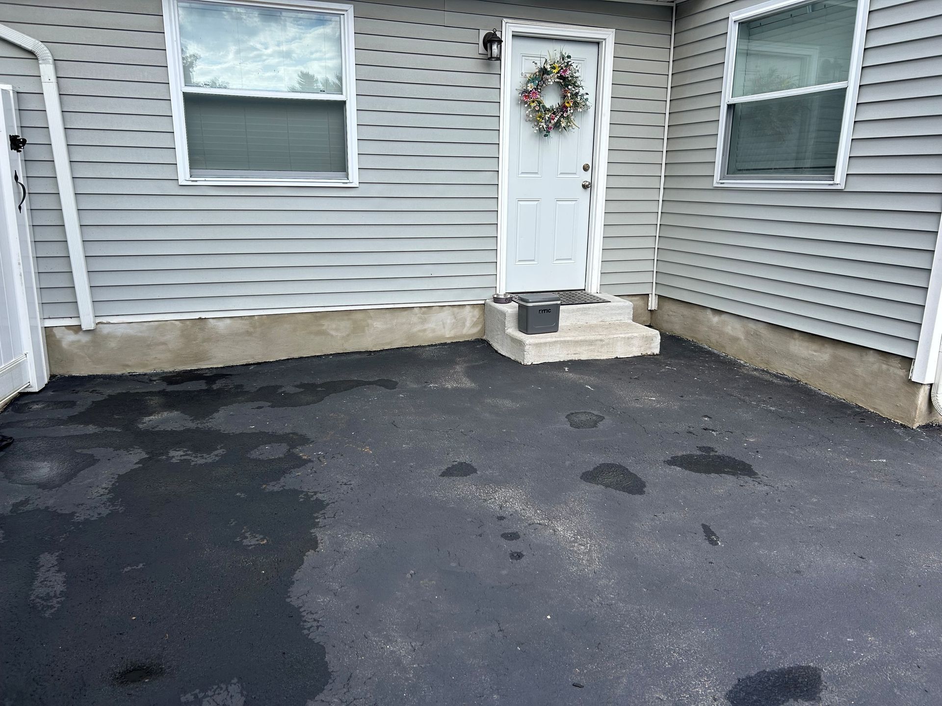 Asphalt driveway with water spots in front of a light gray house with a wreath on the front door.