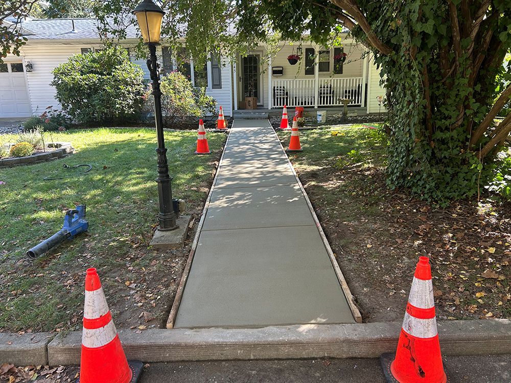 Newly poured concrete sidewalk leading to a house, flanked by orange traffic cones.