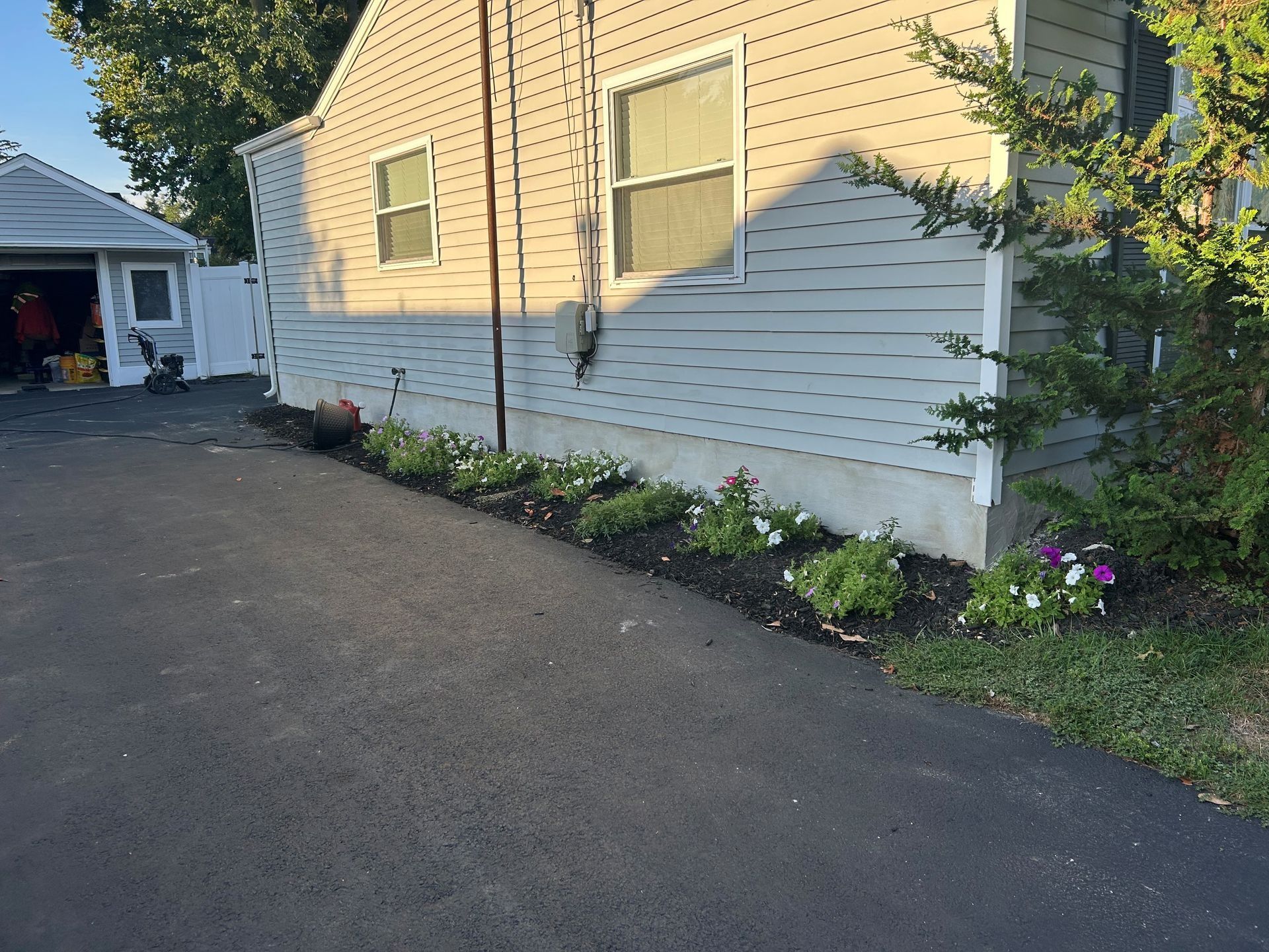 Asphalt driveway next to a house with a flower bed. Sunlight illuminates the building.
