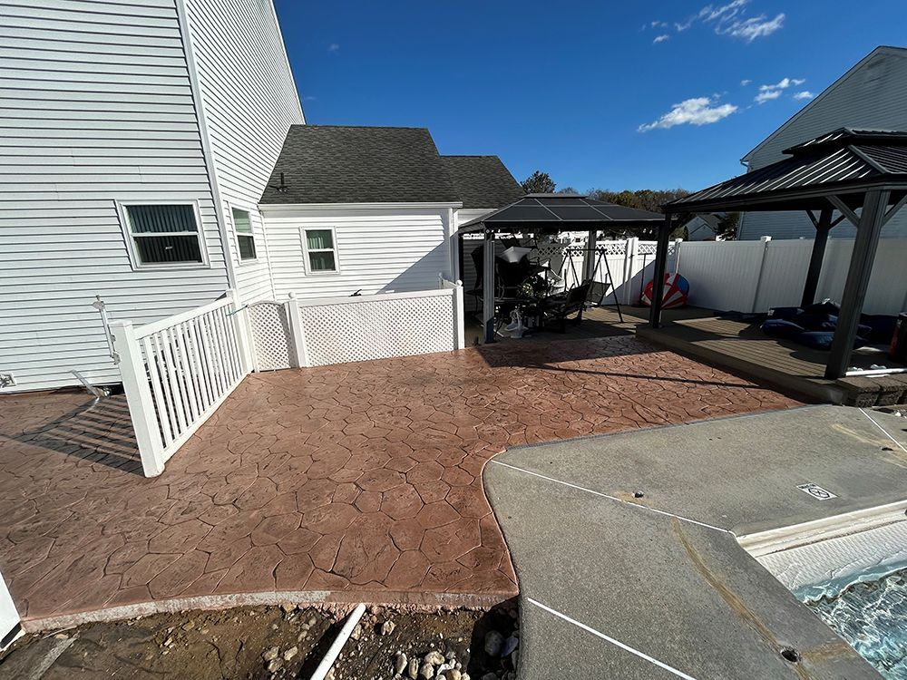 Brown stamped concrete patio next to a white fence and pool. A gazebo and small building are in the background.
