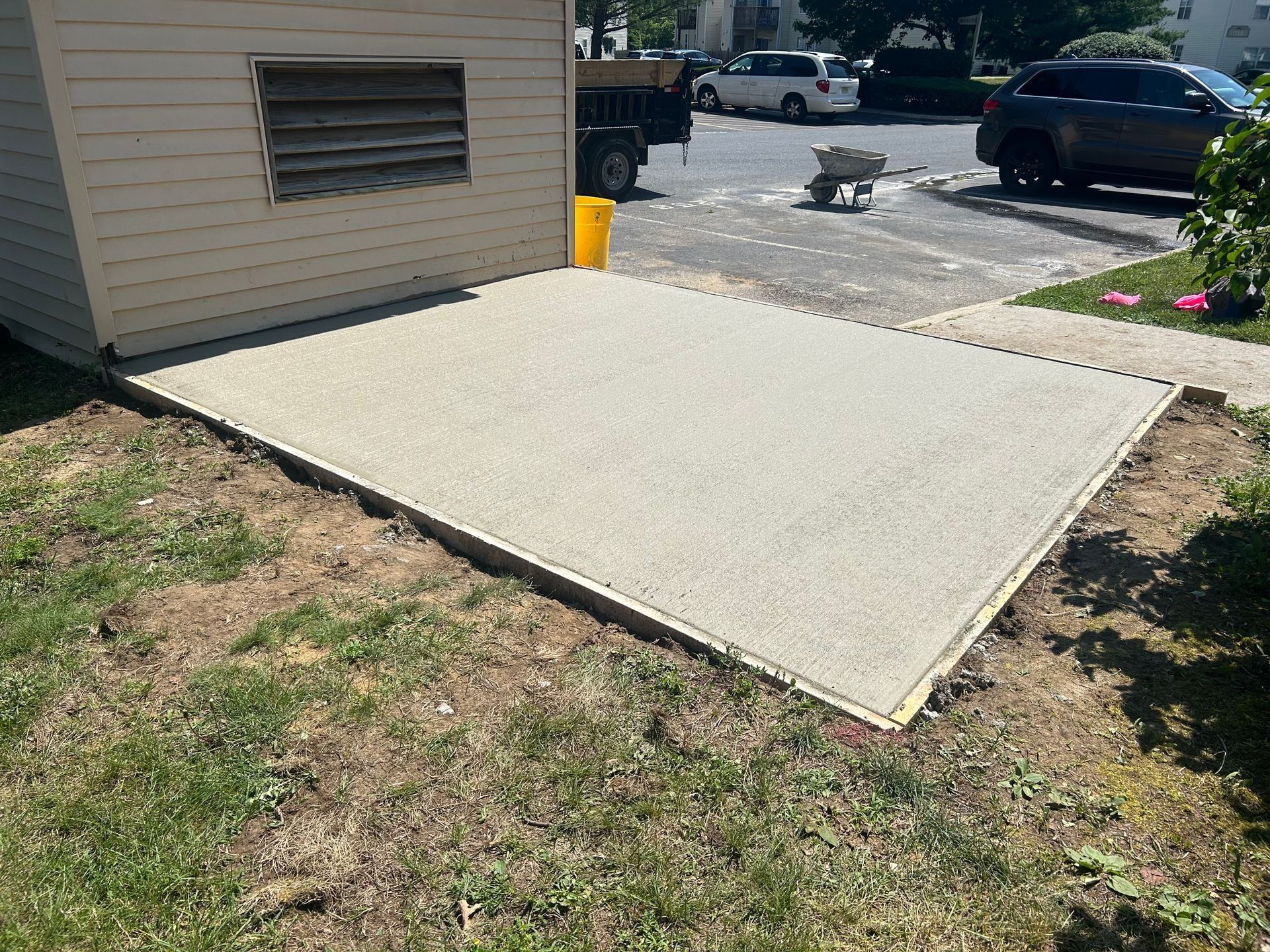 Newly poured concrete patio next to a shed, surrounded by grass and sidewalk.