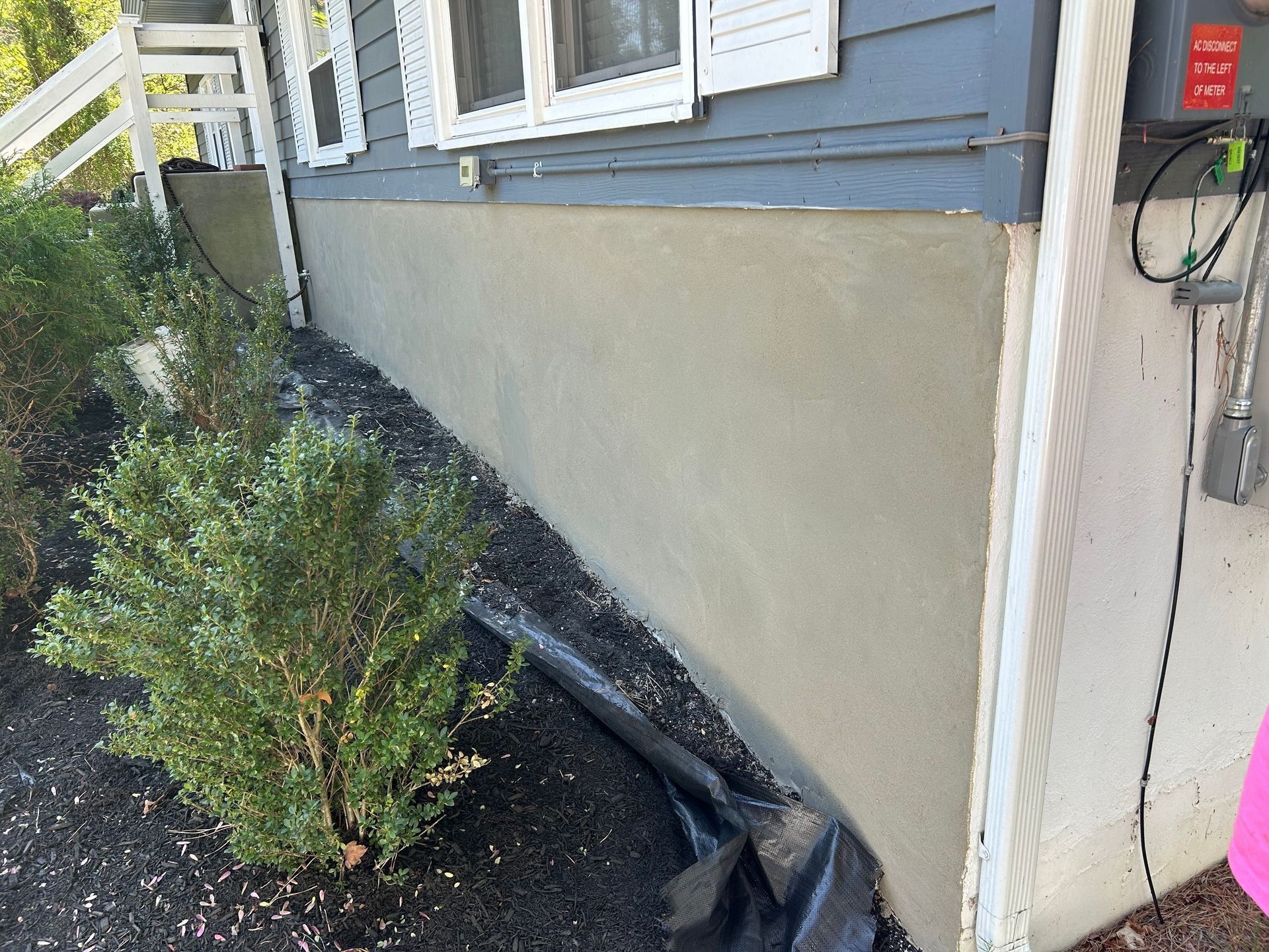 Gray stucco foundation on a house, with bushes, black mulch, and a utility box visible.