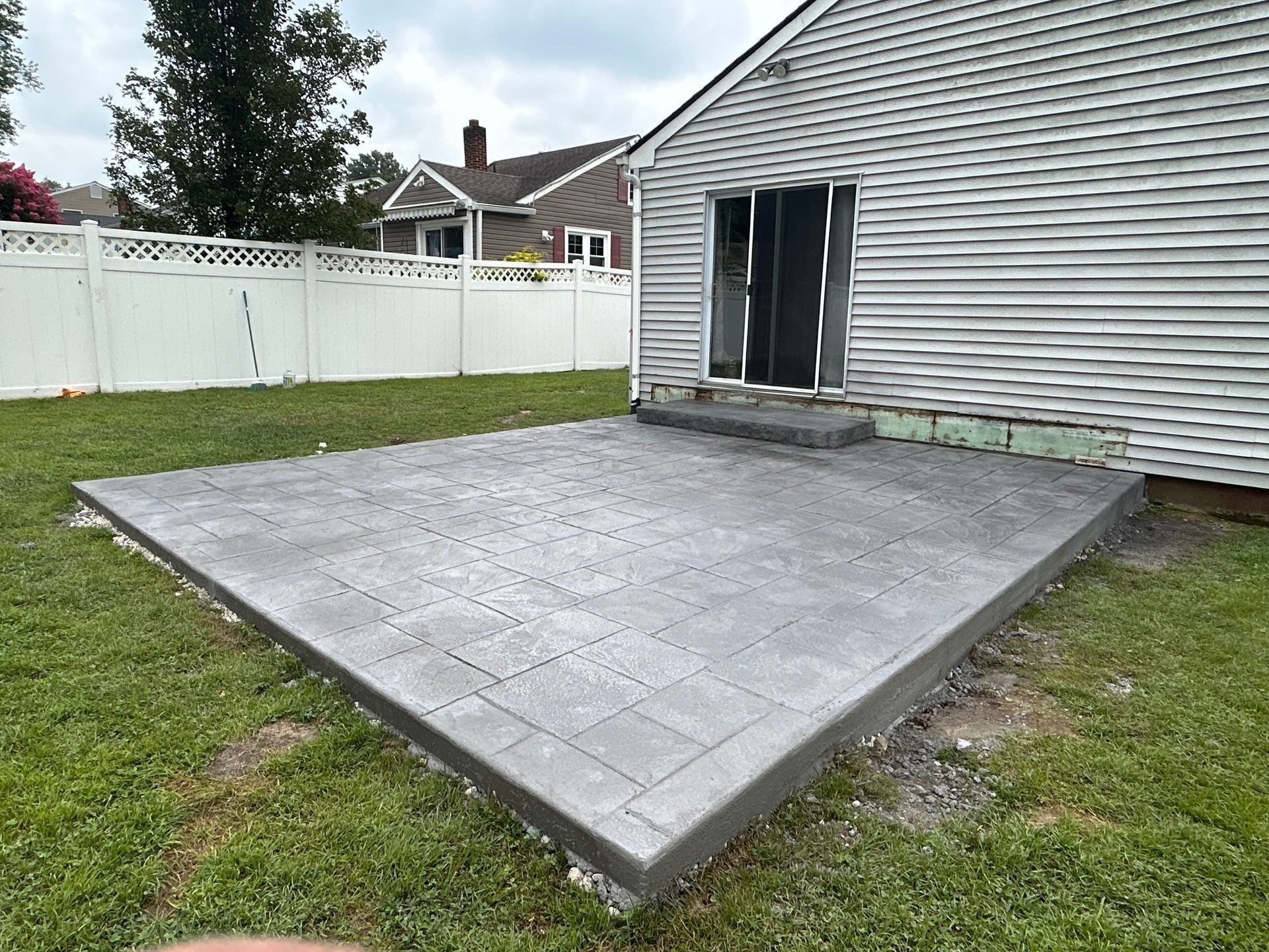 Concrete patio next to a house with sliding glass door, surrounded by grass and a white fence.