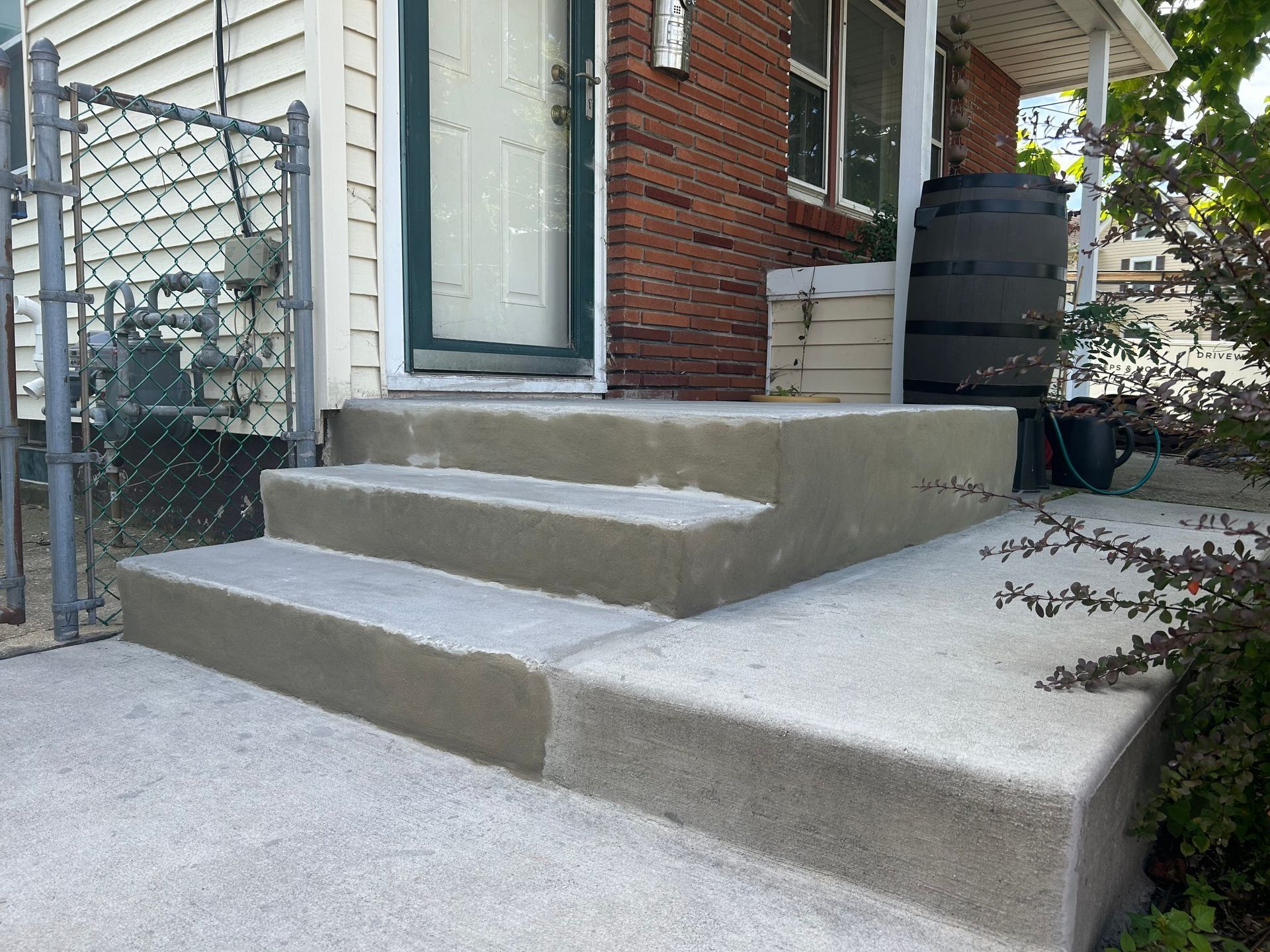 Concrete steps leading to a house's front door. A gray chain-link fence and brick facade are also visible.