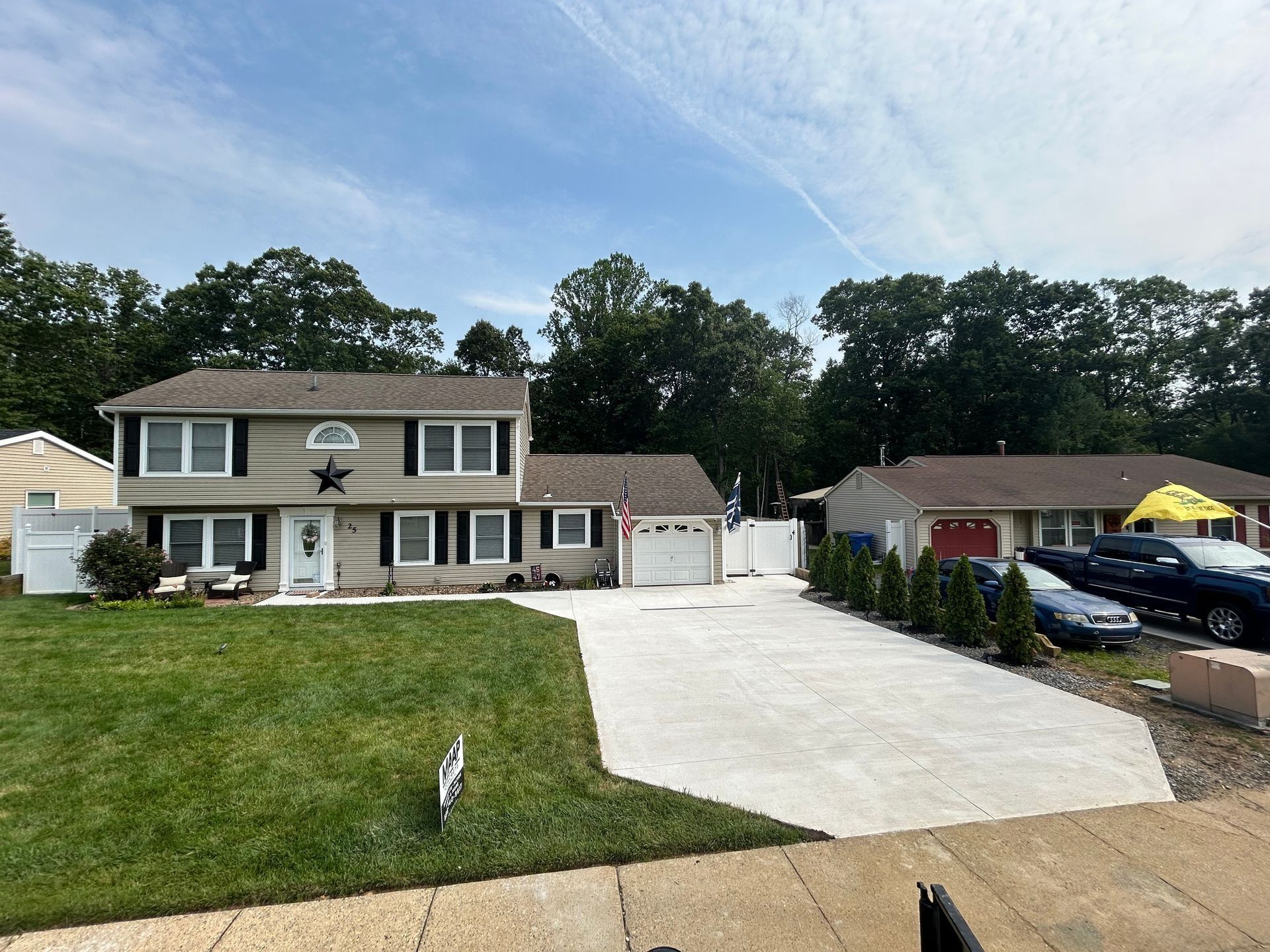 Beige house with black shutters, a wide concrete driveway, and a green lawn on a sunny day.