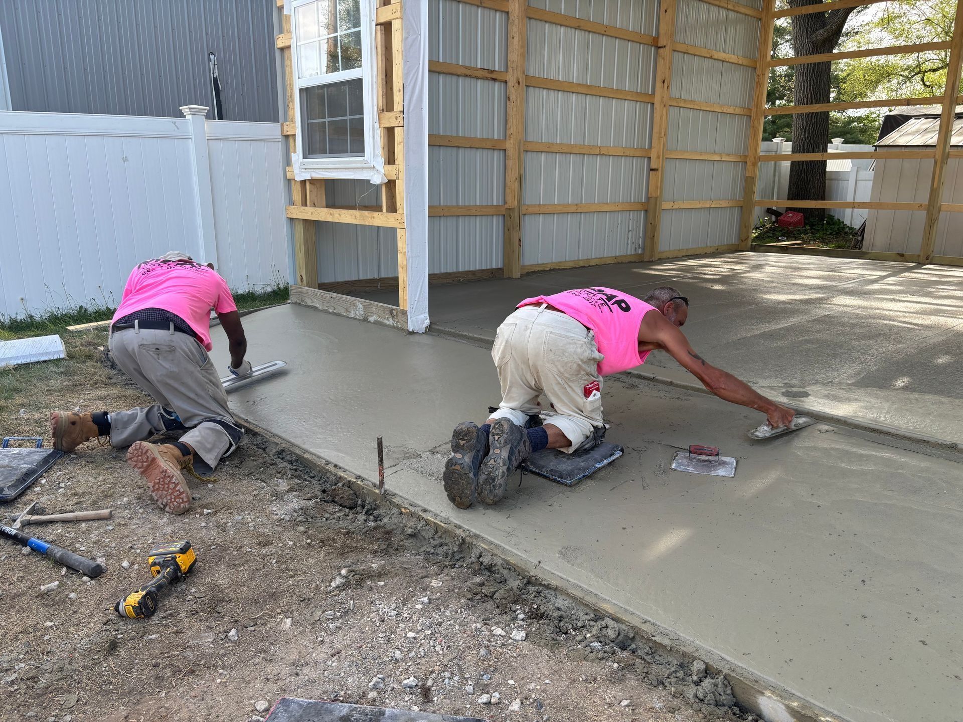 Two workers smoothing wet concrete on a construction site, wearing pink shirts and knee pads.