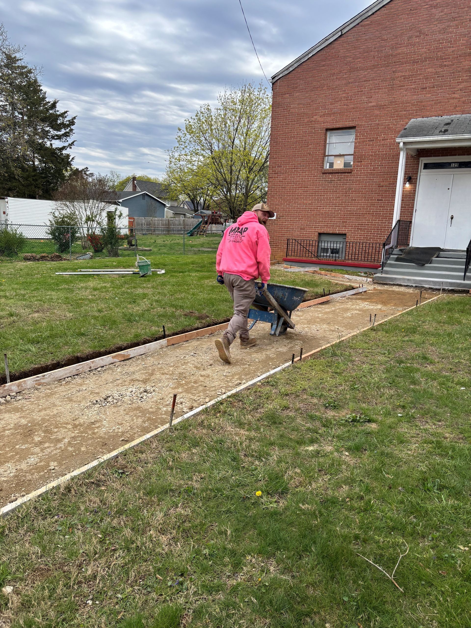 Man in pink shirt pushes a wheelbarrow along a gravel pathway being built next to a building.