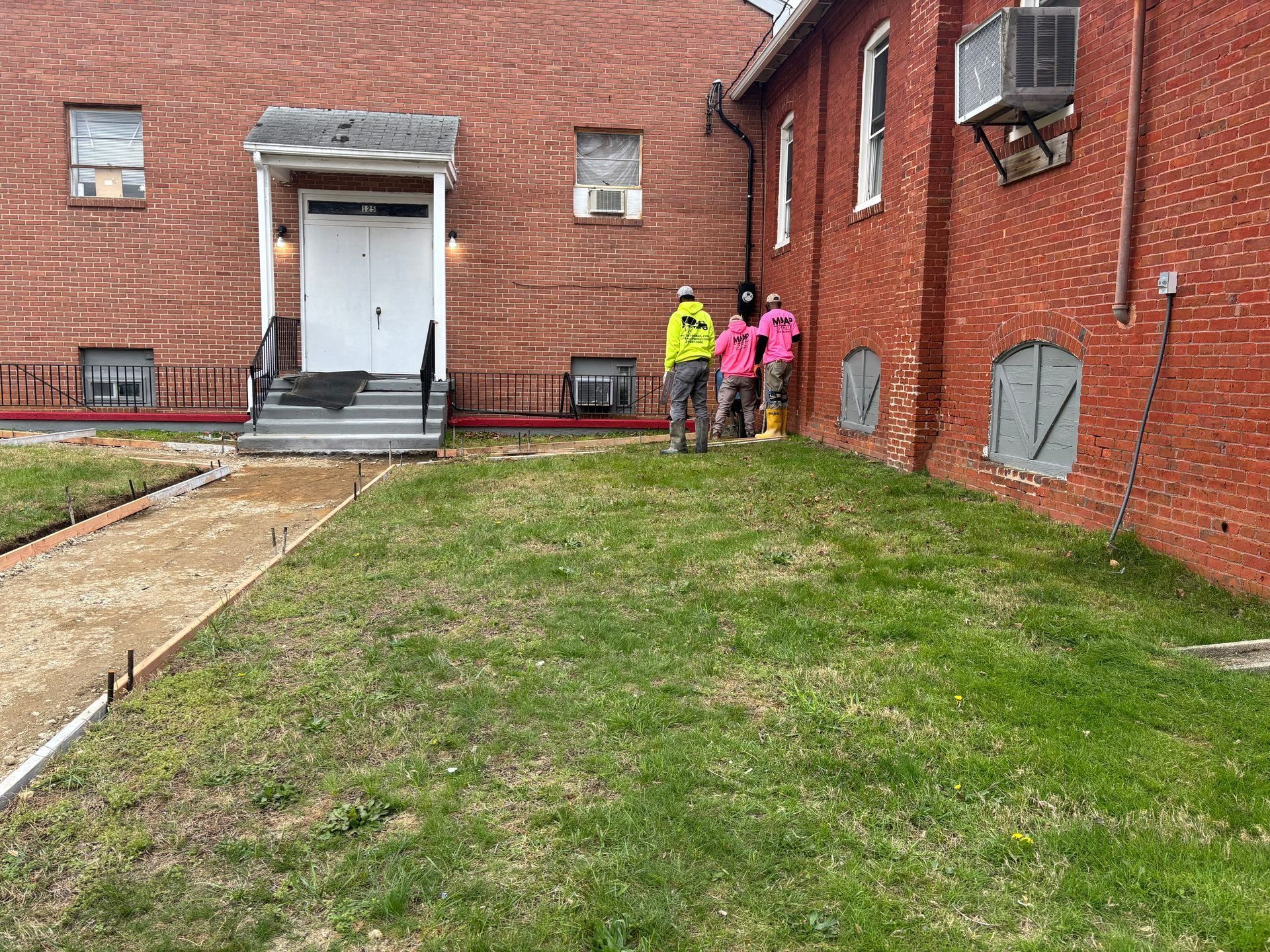 Workers in bright vests stand near a brick building with an overgrown lawn and unfinished path.