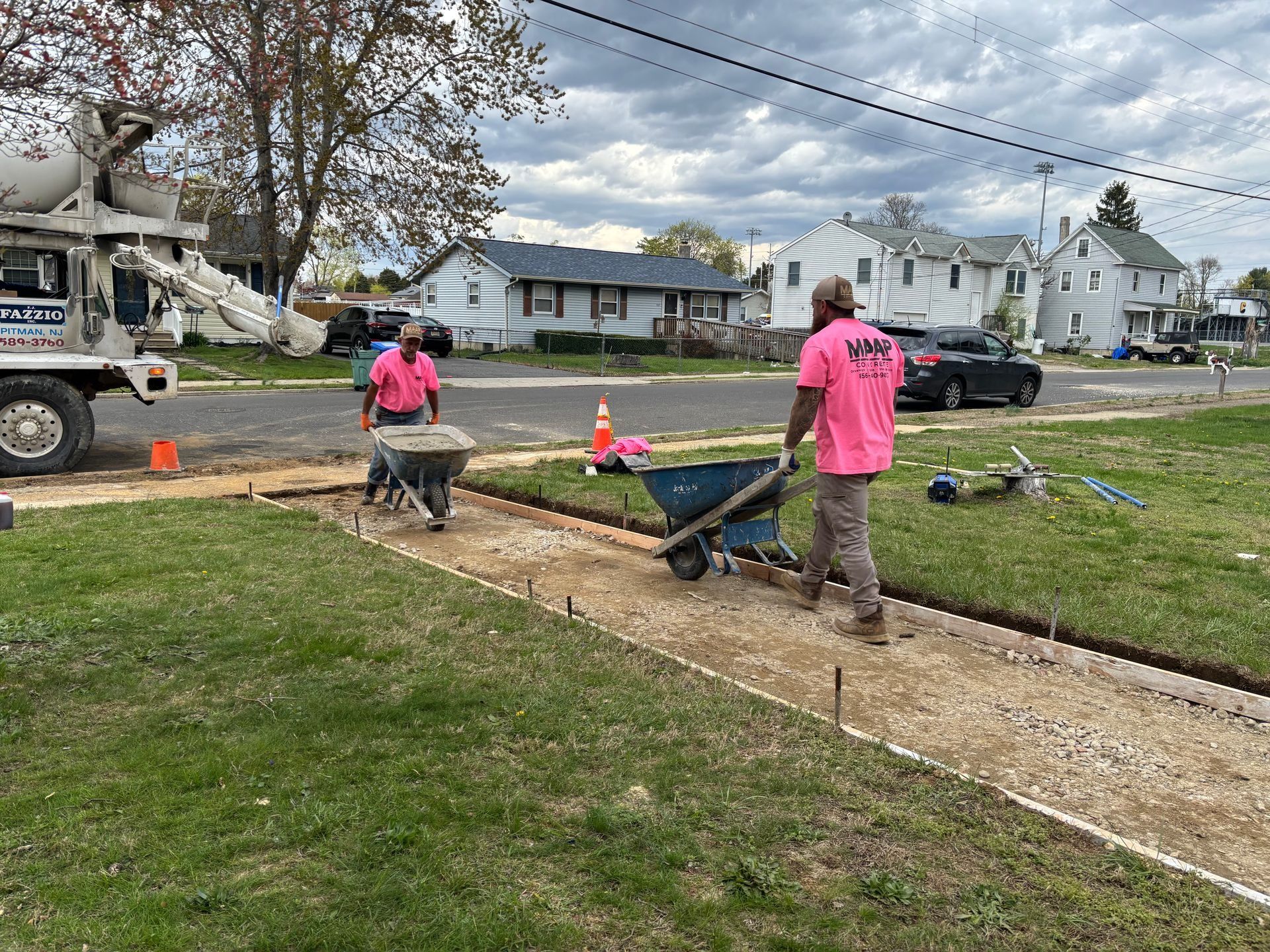 Two workers in pink shirts pour concrete from a truck into a sidewalk form on a residential street.