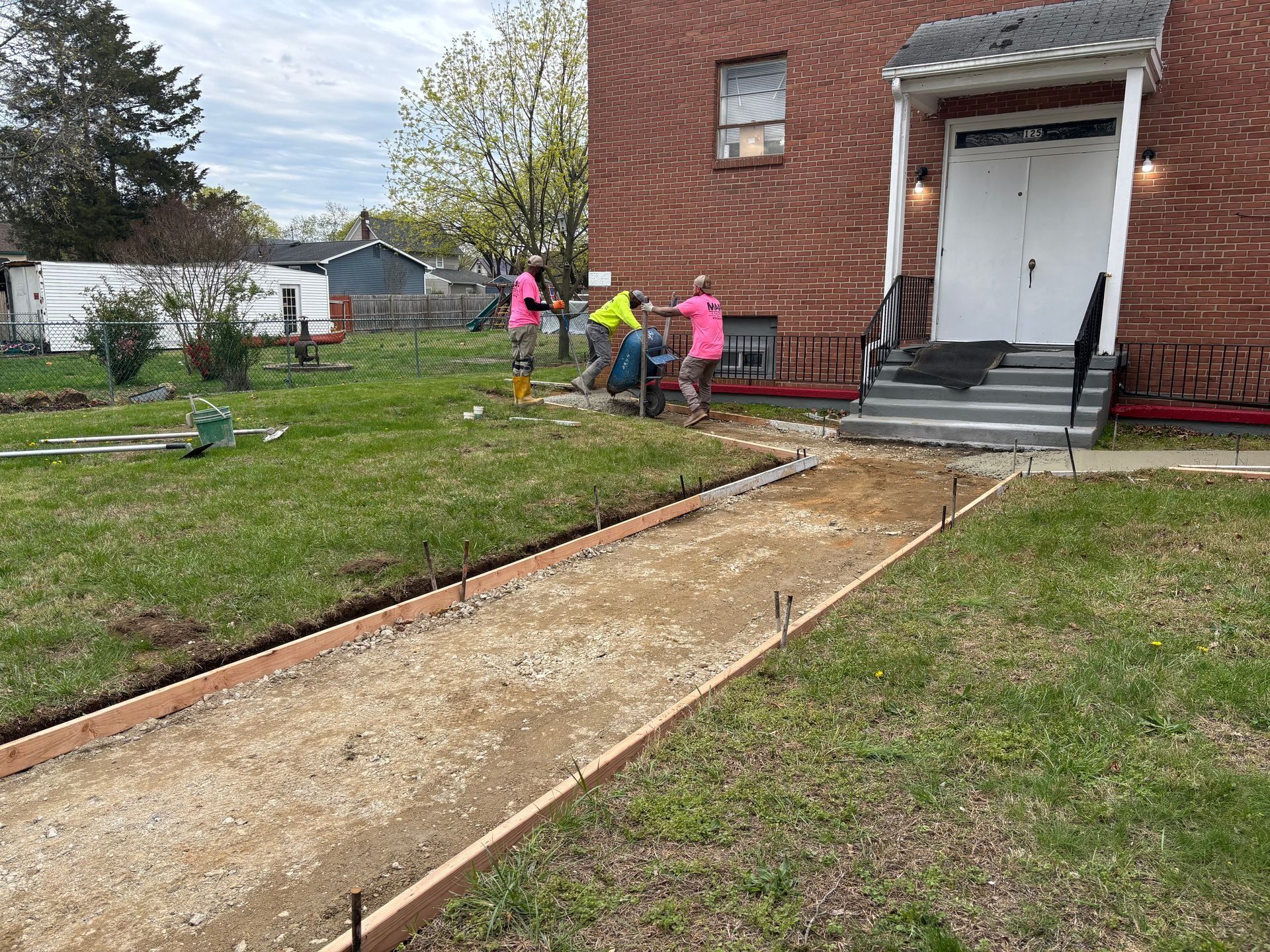 Workers constructing a concrete pathway in front of a brick building, using pink and yellow safety gear.