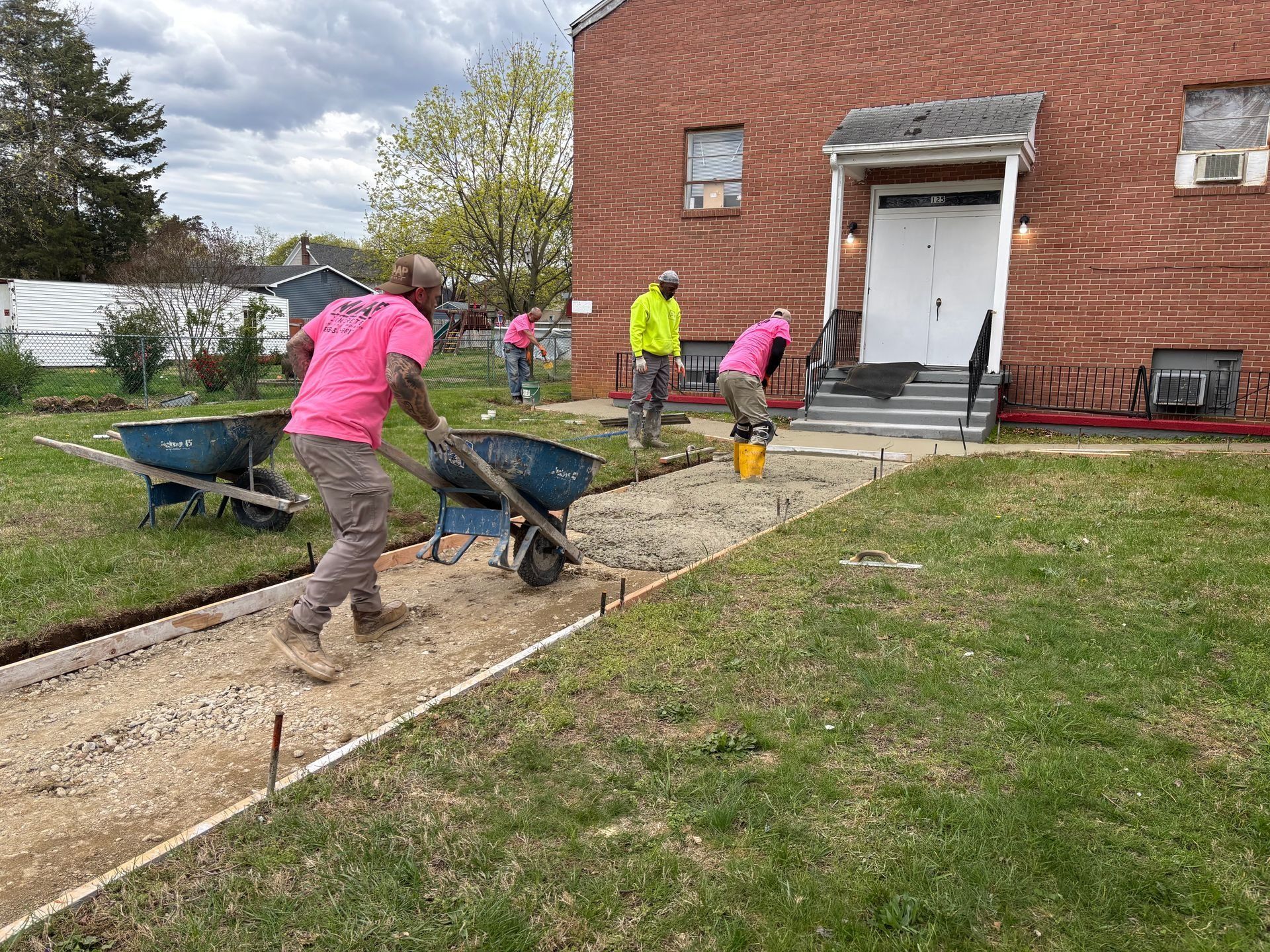 Construction workers in pink shirts pour gravel for a sidewalk near a red brick building.