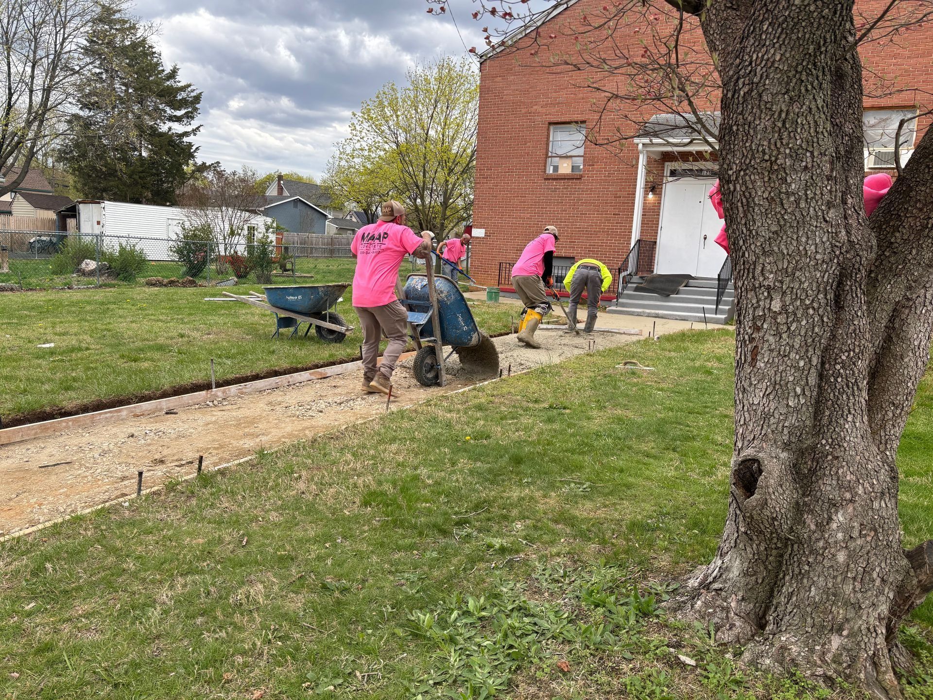 People in pink shirts working on a walkway outside a brick building; wheelbarrows and cement mixer present.