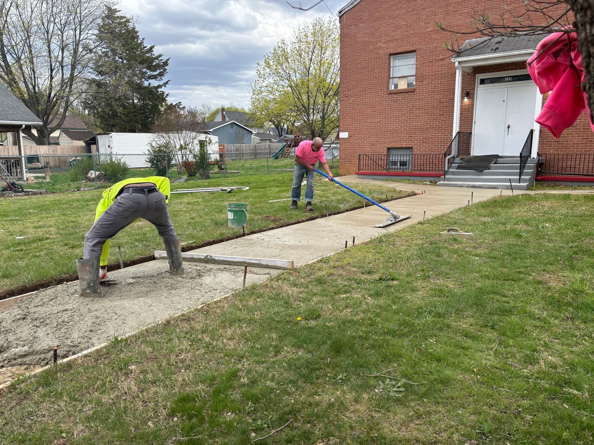 Workers pouring and leveling concrete on a walkway in front of a brick building.