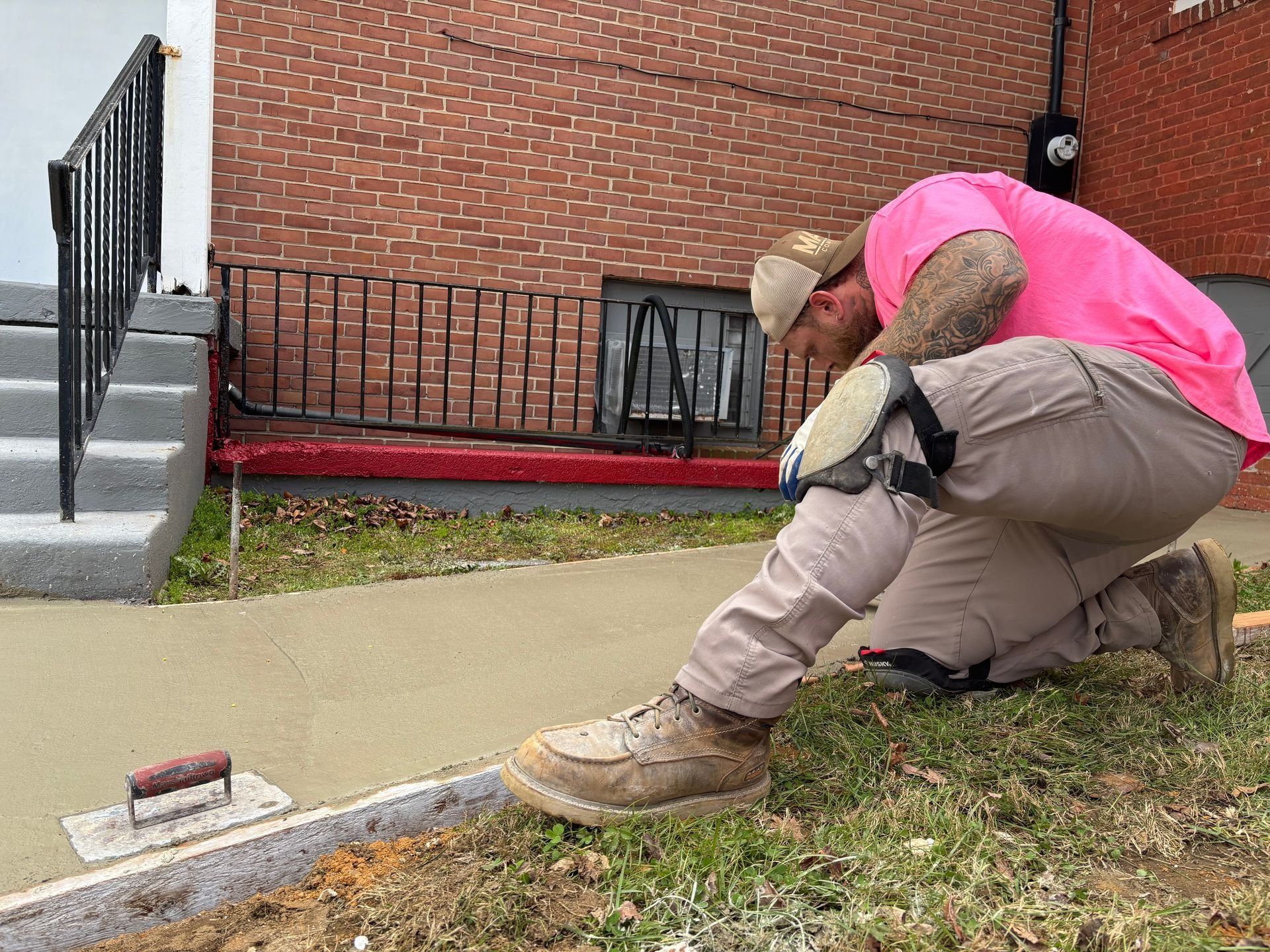 Man in pink shirt and knee pads finishing concrete sidewalk in front of a brick building.