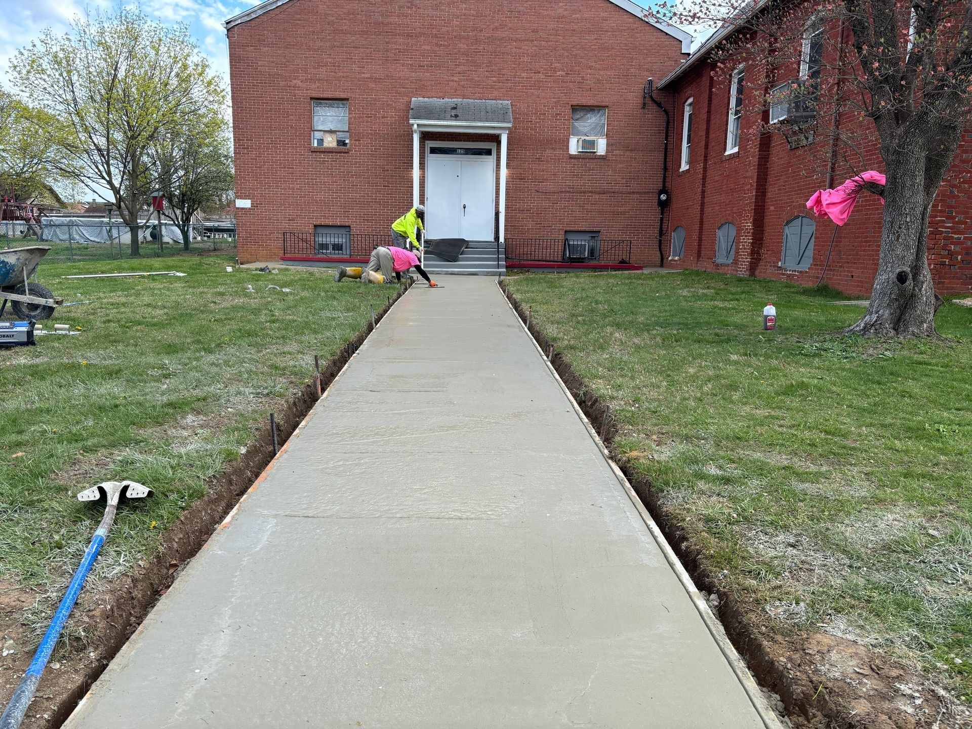 Workers pouring concrete sidewalk in front of a brick building. Green lawn borders the new path.