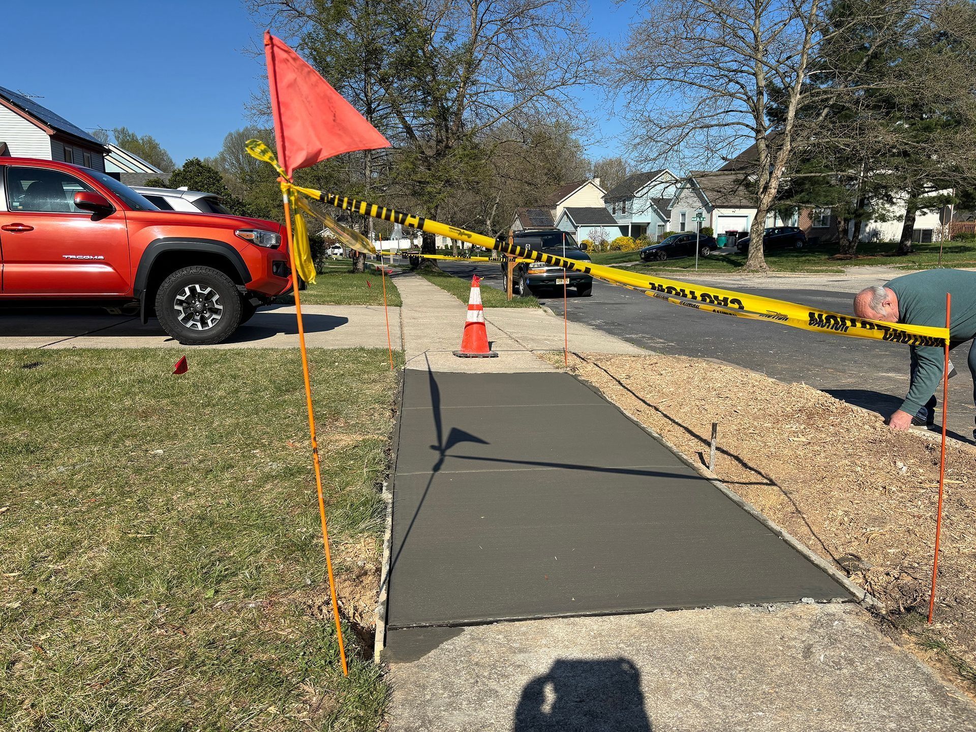 Concrete walkway being poured; caution tape, orange flag, and truck in the background.