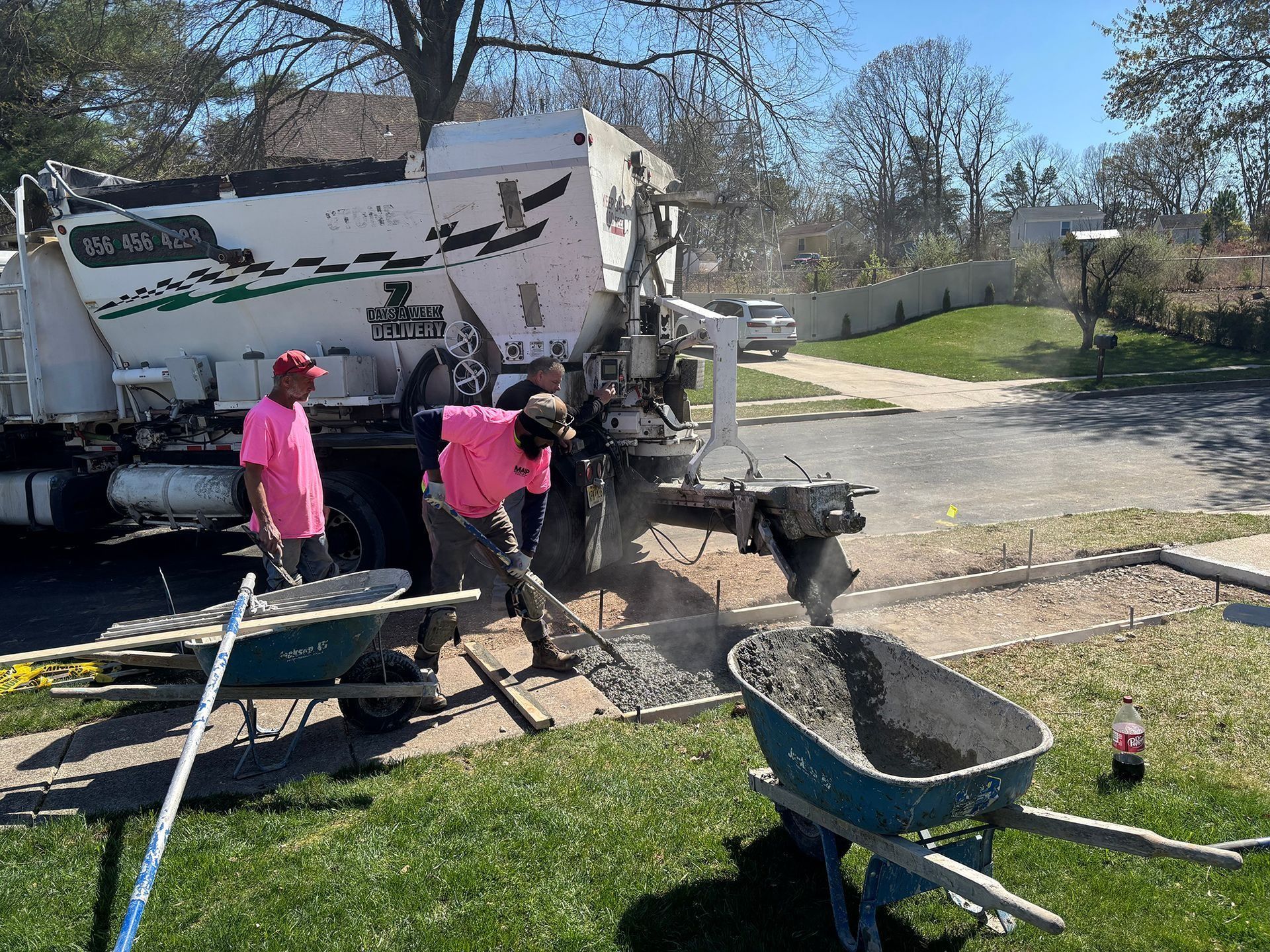 Men in pink shirts pouring concrete from truck into wheelbarrow on a lawn.