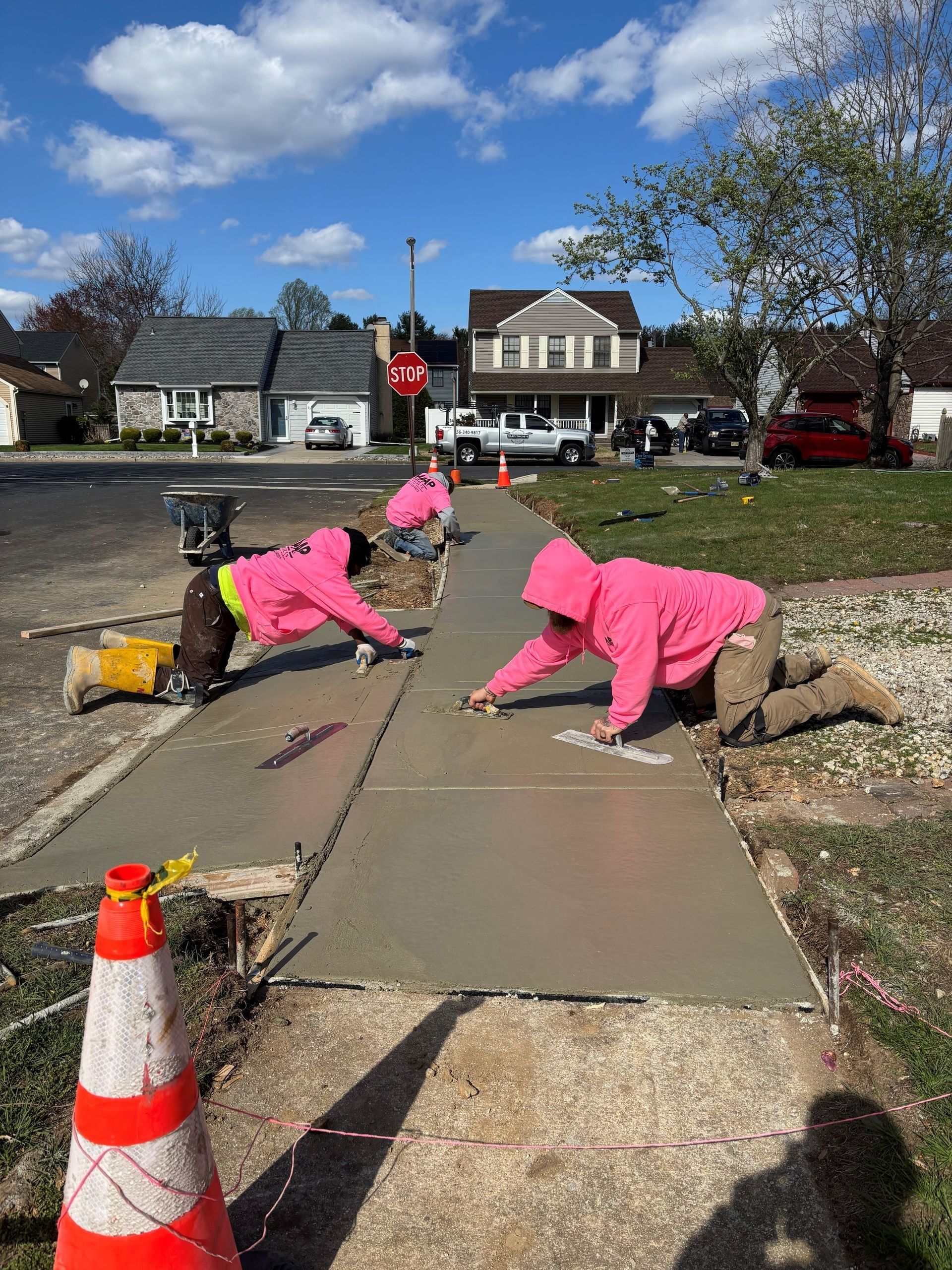 Three workers in pink hoodies smoothing wet cement on a sidewalk in front of houses on a sunny day.