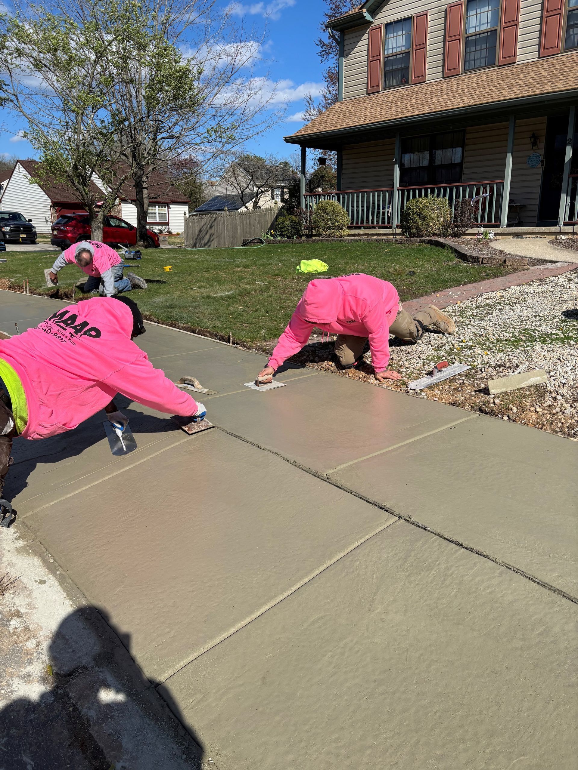 Three workers in pink coveralls smoothing fresh concrete sidewalk outside a house on a sunny day.