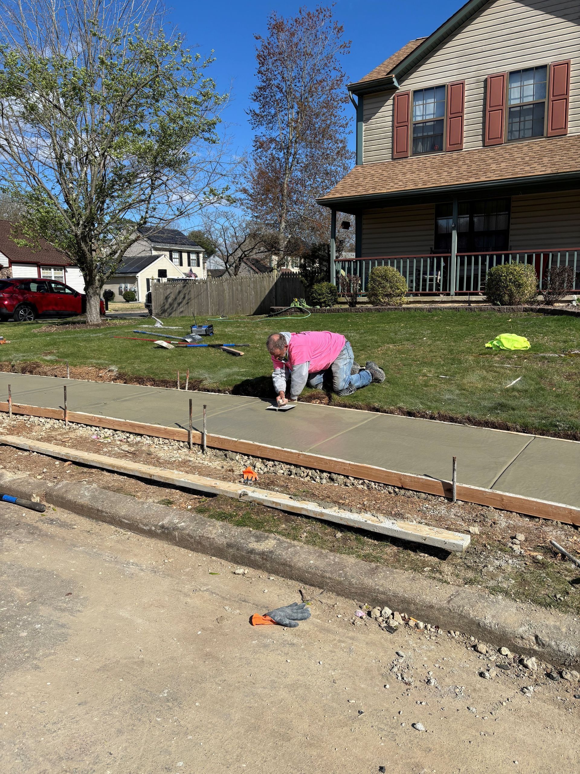 Person kneeling on grass, smoothing wet concrete sidewalk. House in background, sunny day.