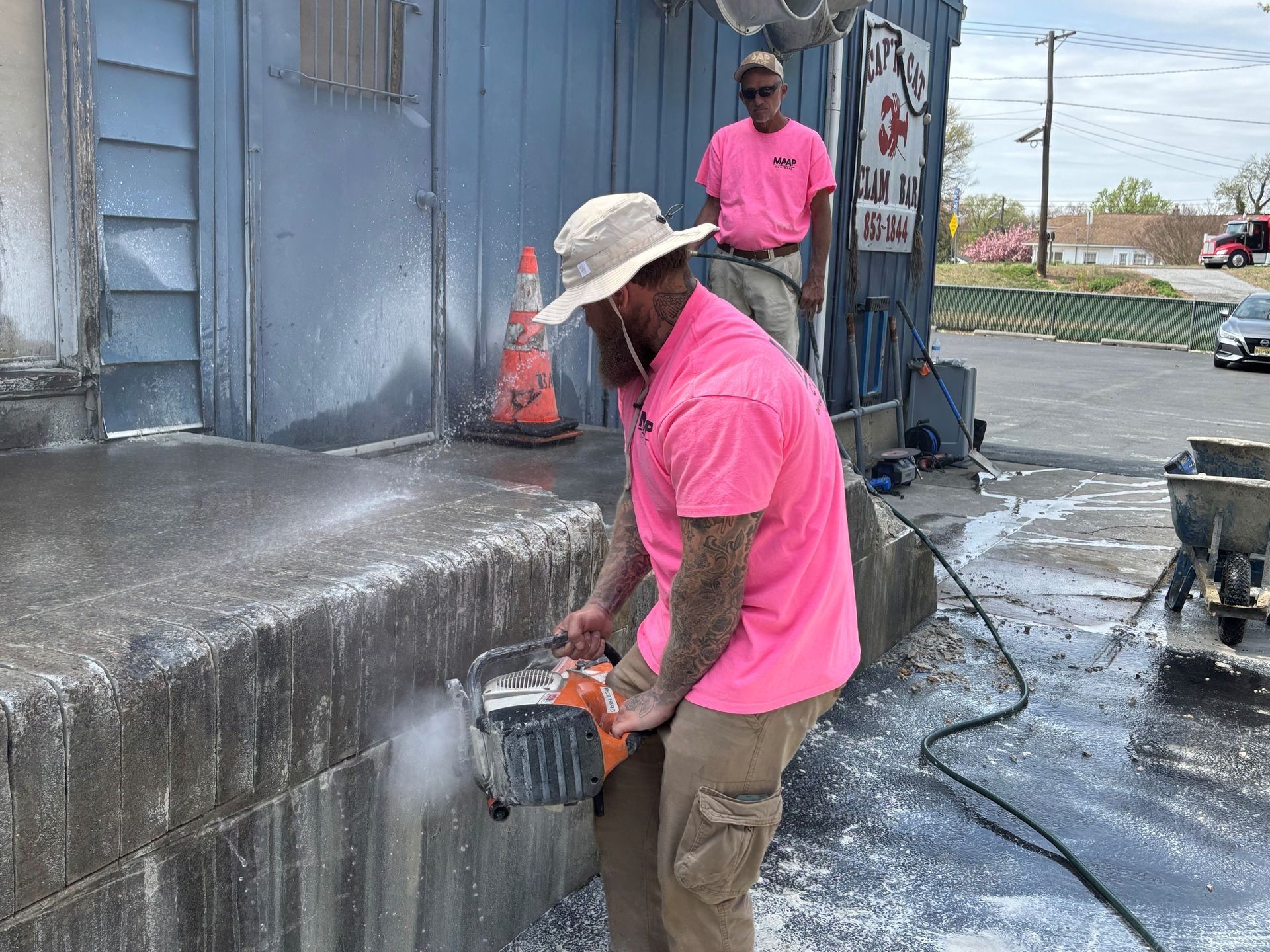 Two men in pink shirts use a saw and water to cut concrete steps outside a blue building.