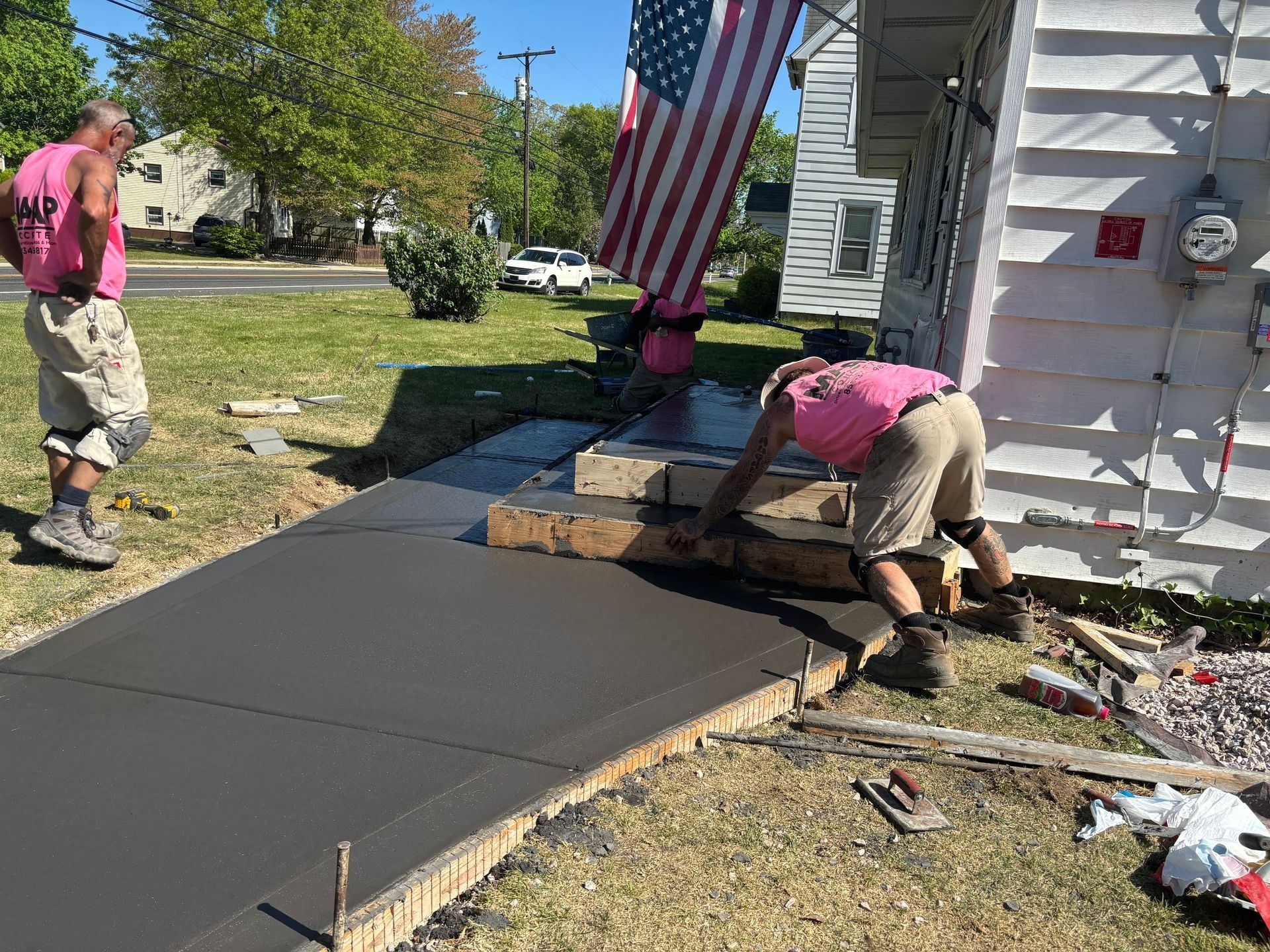 Three construction workers pouring and smoothing concrete walkway near a house. One holds an American flag.