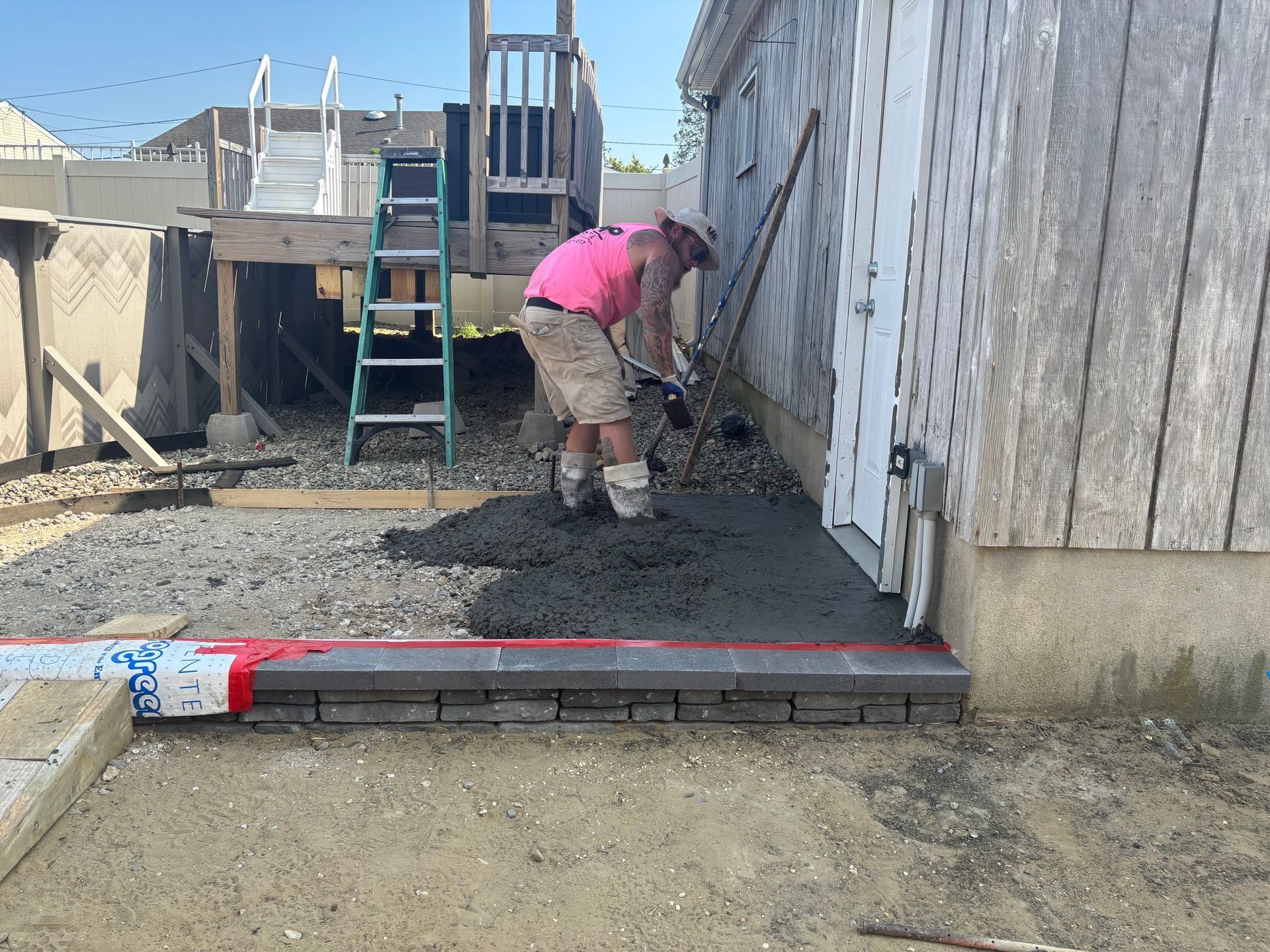 Man pouring concrete next to building entrance; brick border, gravel, and wooden deck visible.