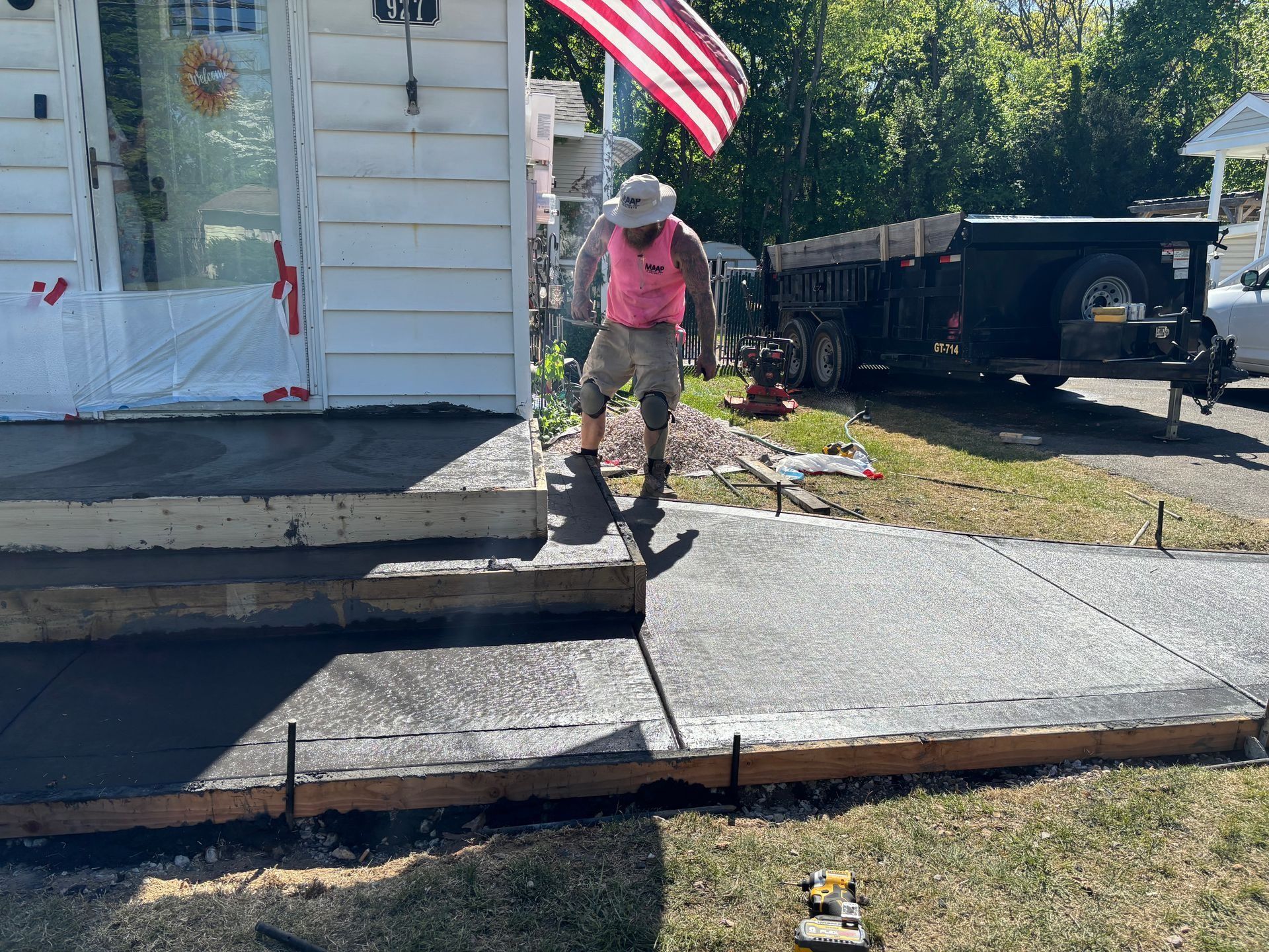 Man in pink shirt walks on a newly poured concrete walkway and steps in front of a house with an American flag.