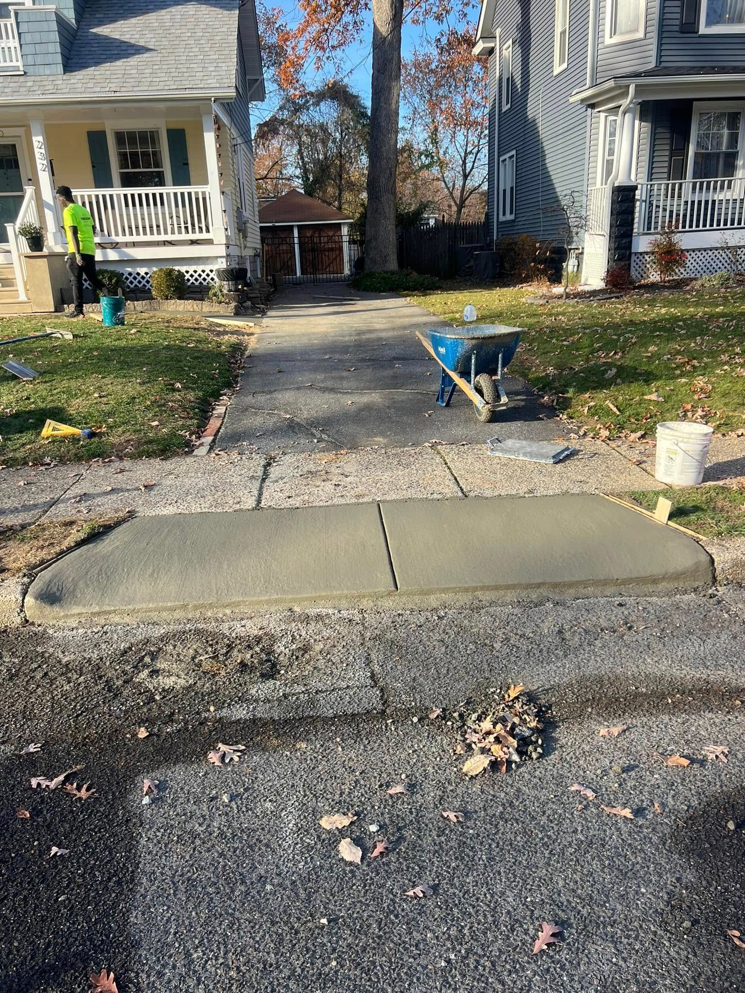 Newly poured concrete walkway section in front of a house. Construction tools and workers present.