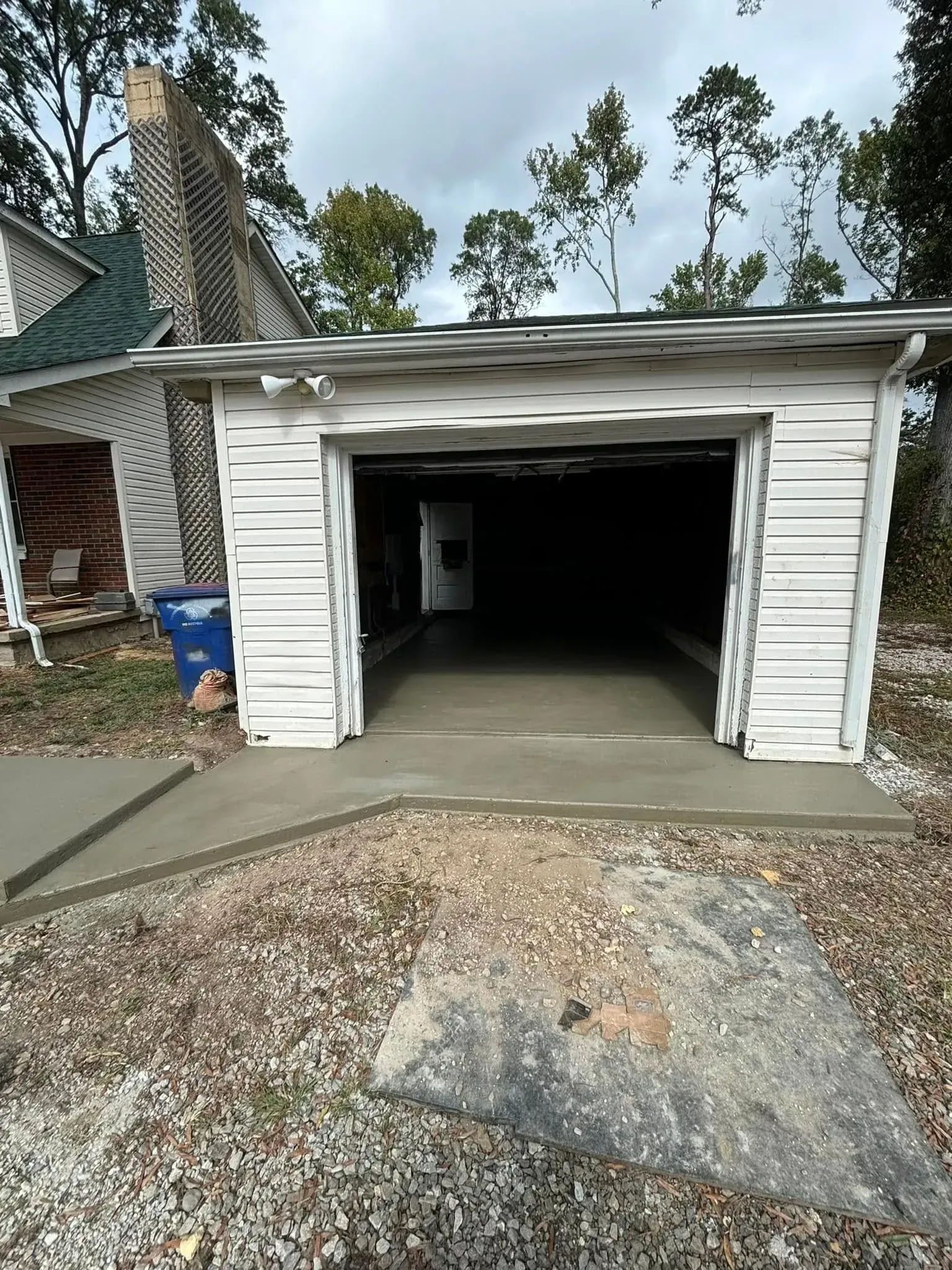 White garage with open door, freshly poured concrete, and a large rock in front. Cloudy sky.