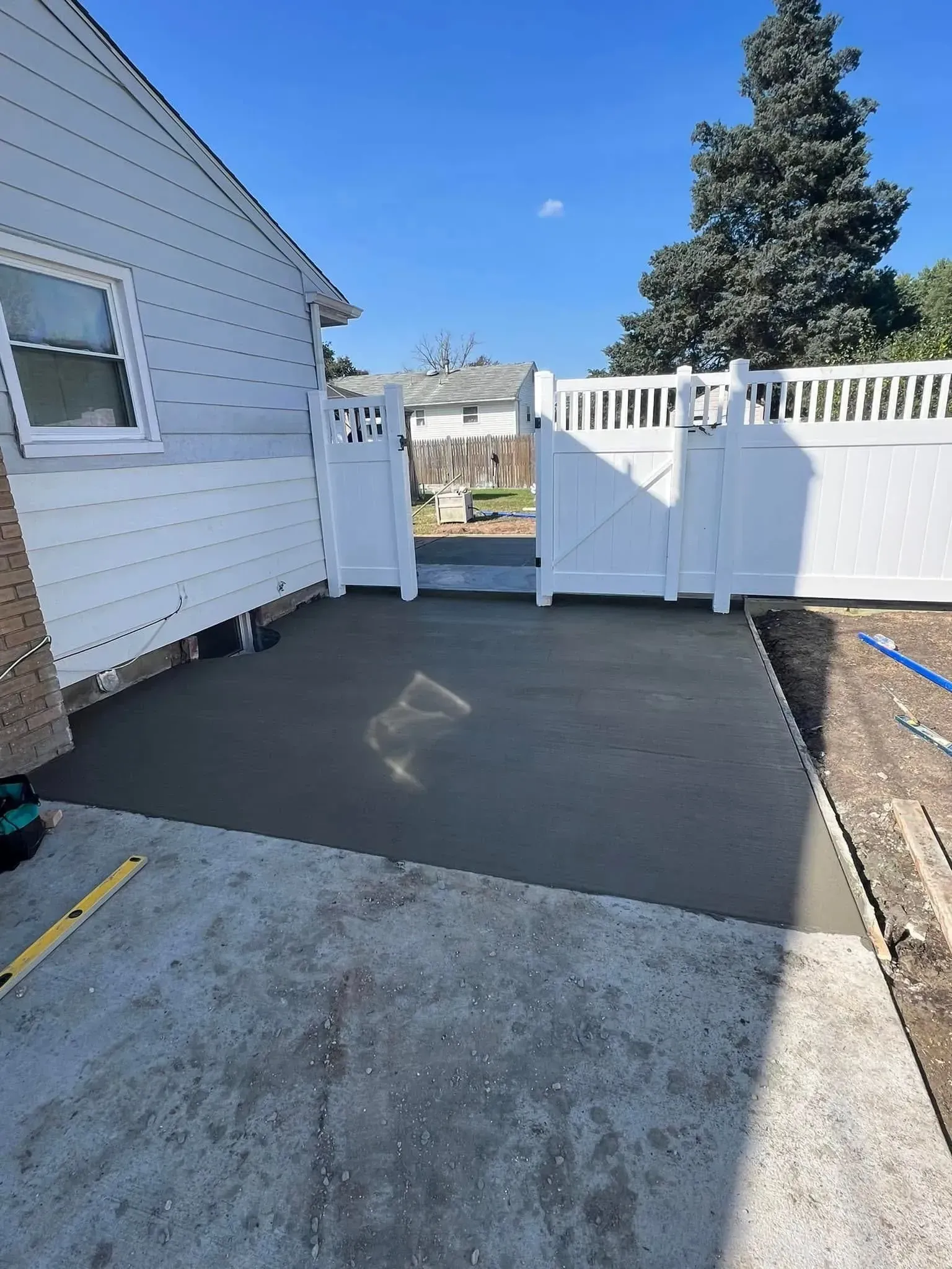 Newly poured concrete patio area with white fence and gate, beside a light-colored house under a blue sky.