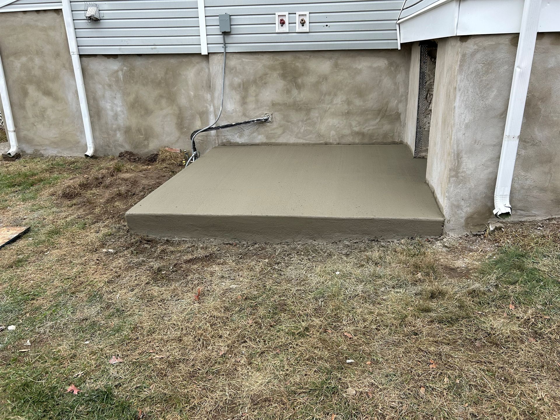 New concrete slab at the base of a house. It’s surrounded by dead grass and abuts a stucco foundation.