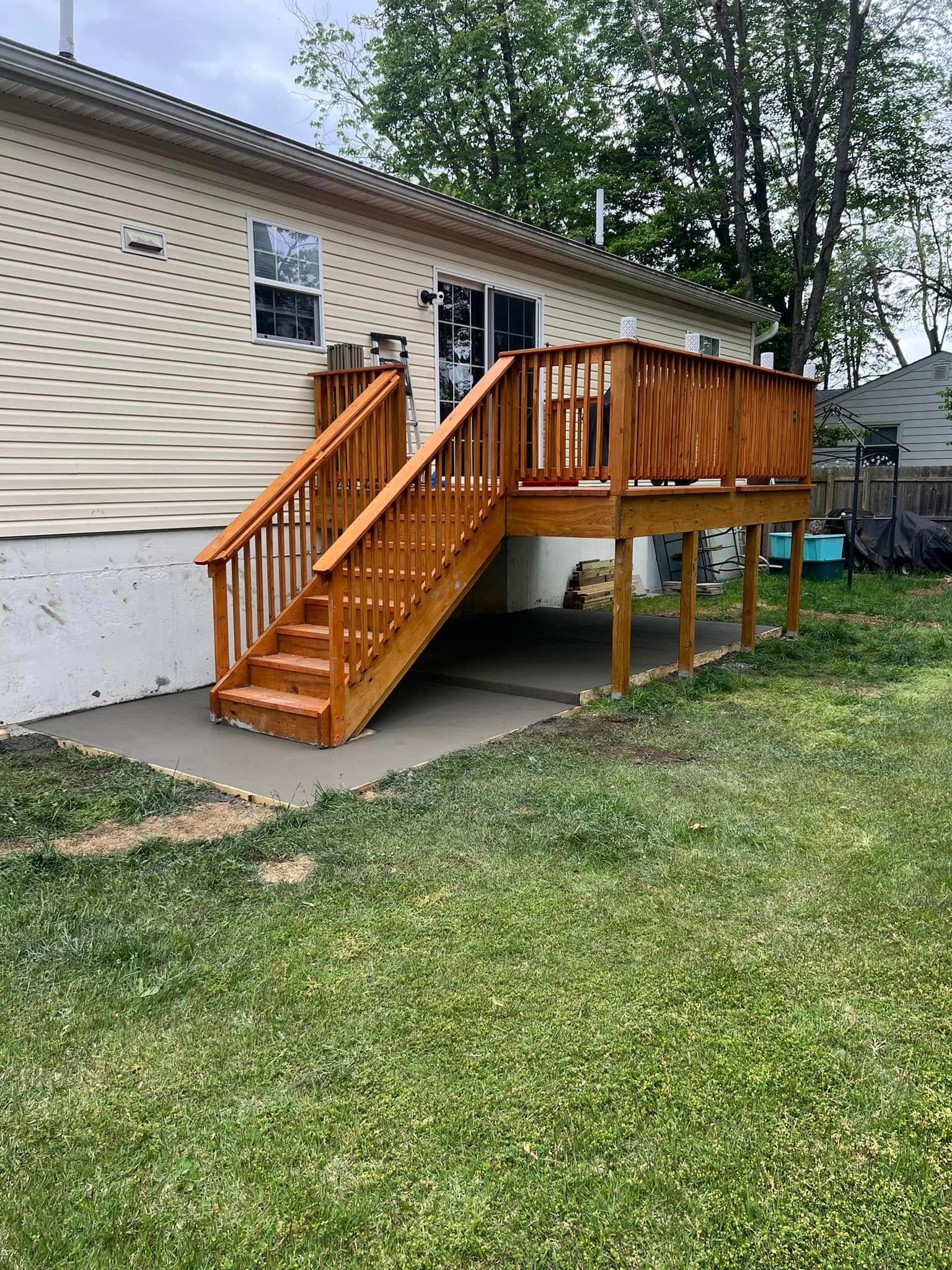 Wooden deck with stairs attached to a tan-sided house. Green grass surrounds a concrete pad beneath the deck.