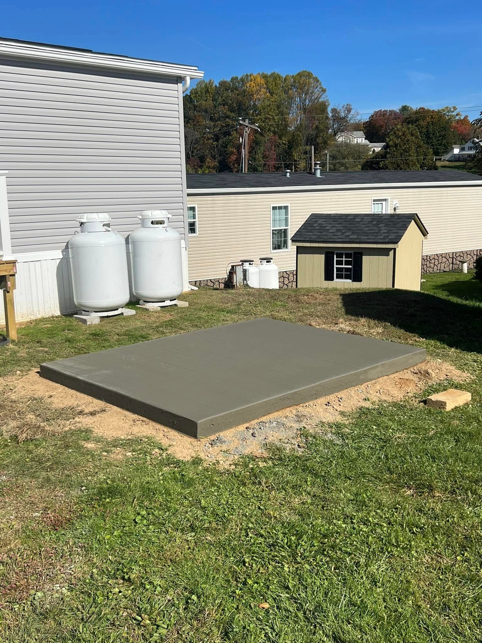 Concrete pad in a yard. Two propane tanks and a small shed are visible nearby. Green grass and a blue sky.