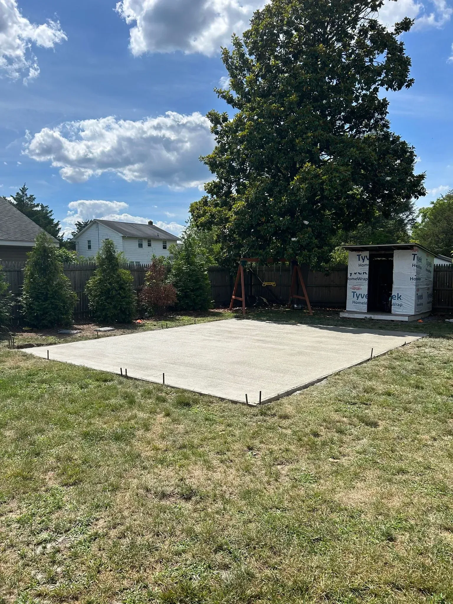 Concrete patio in a backyard, partially built shed, green grass, trees, sunny sky.