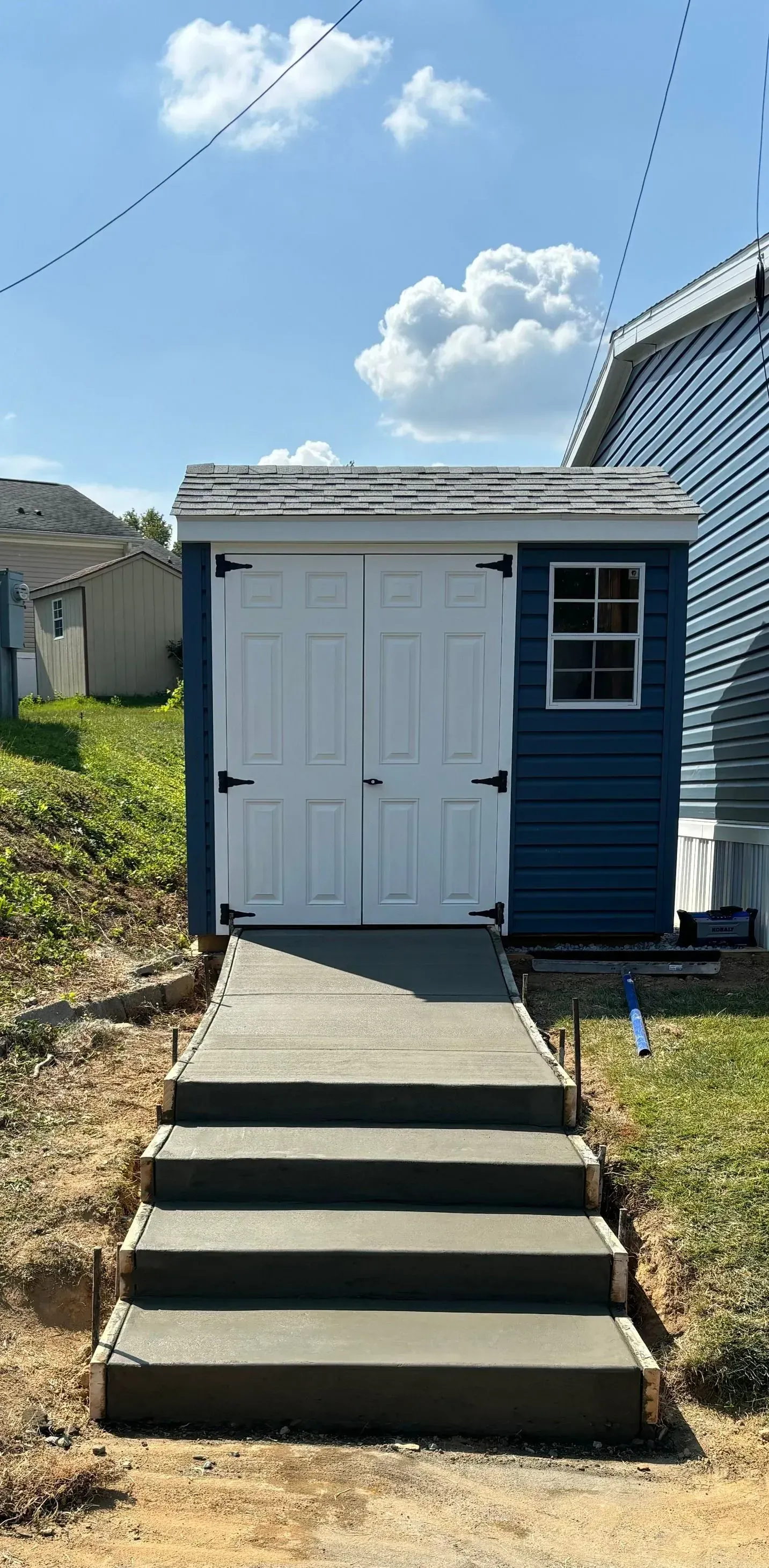 Blue shed with white doors and a concrete path with stairs leading up to it on a sunny day.