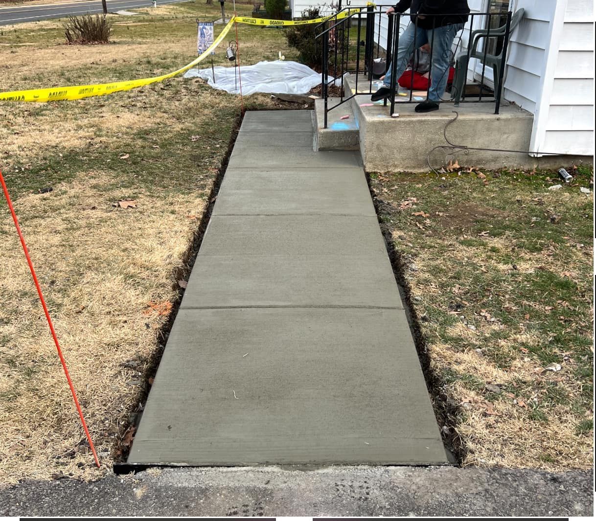 Newly poured concrete sidewalk leading to a house with person standing on porch. Yellow caution tape nearby.