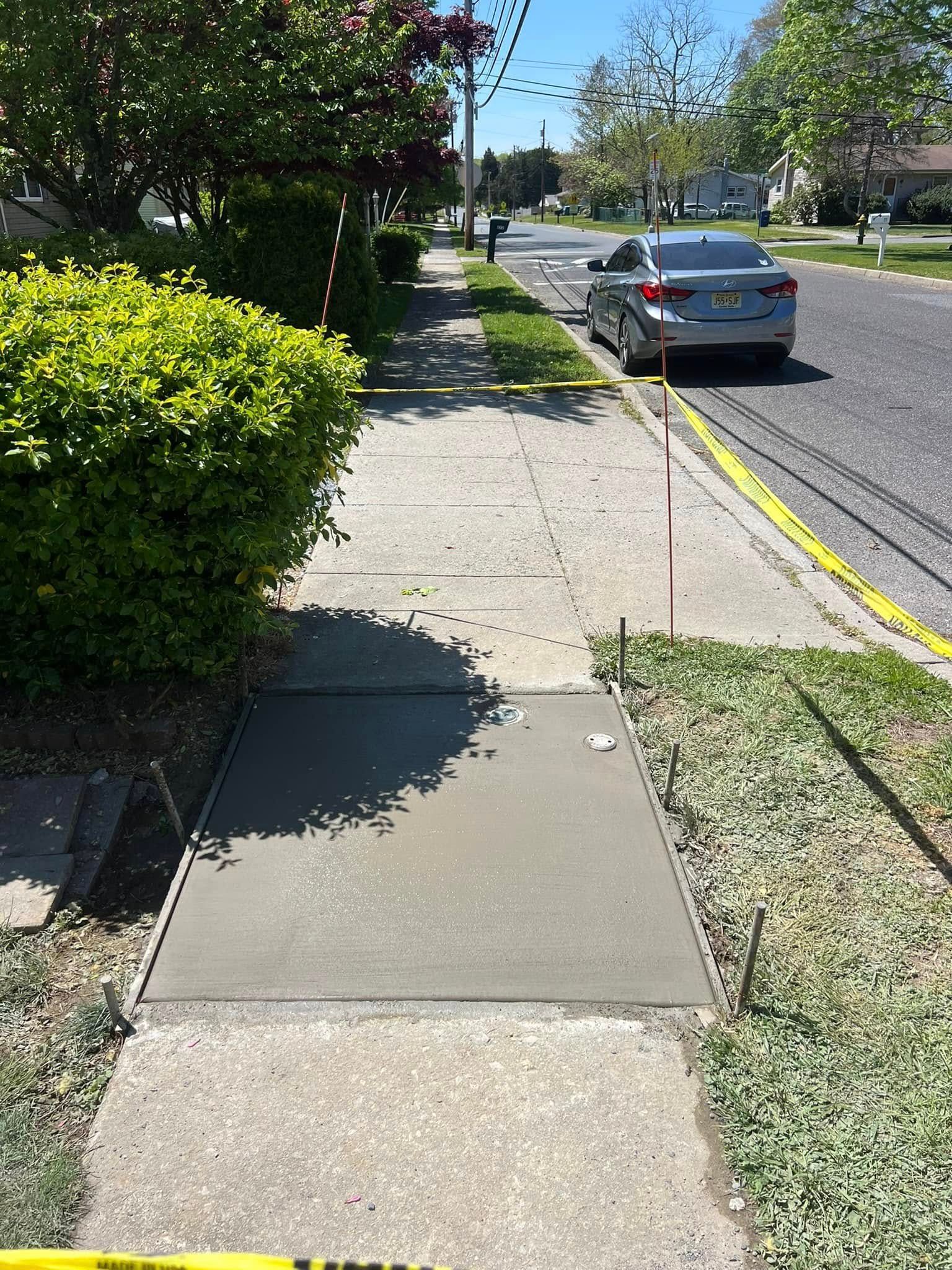 Sidewalk with a newly poured concrete patch, bordered by metal guides, next to a green bush and a street.