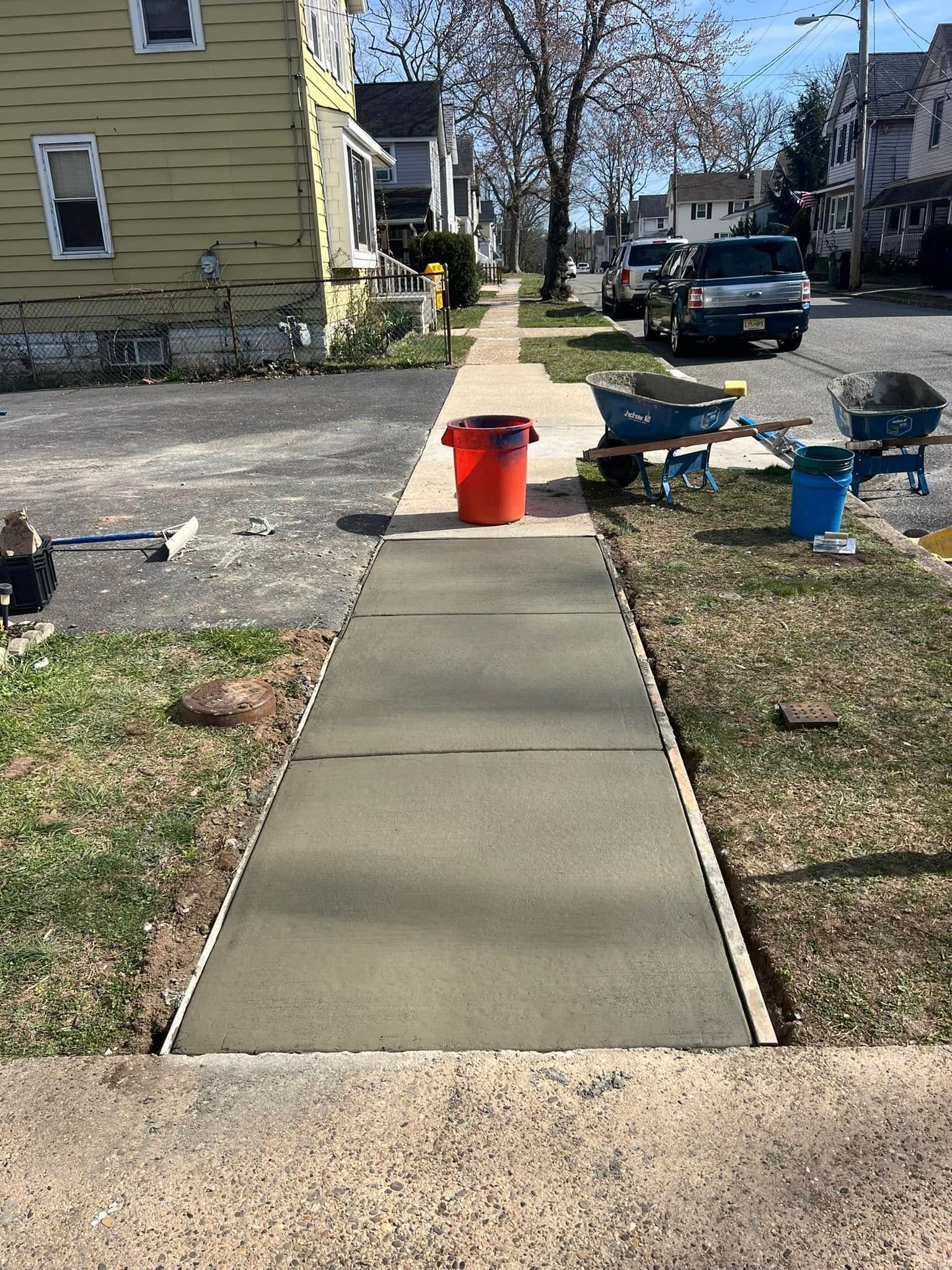 Freshly poured concrete sidewalk with orange bucket, wheelbarrows, and parked vehicles along the street.