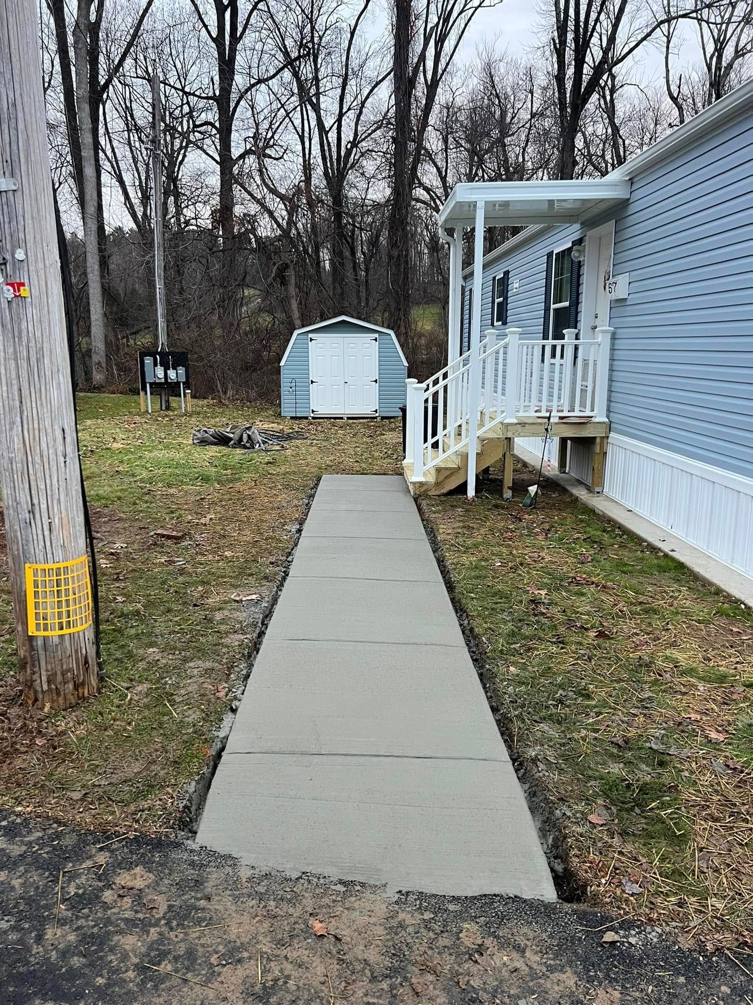 Concrete walkway leading to a blue mobile home's porch, with a shed in the background.