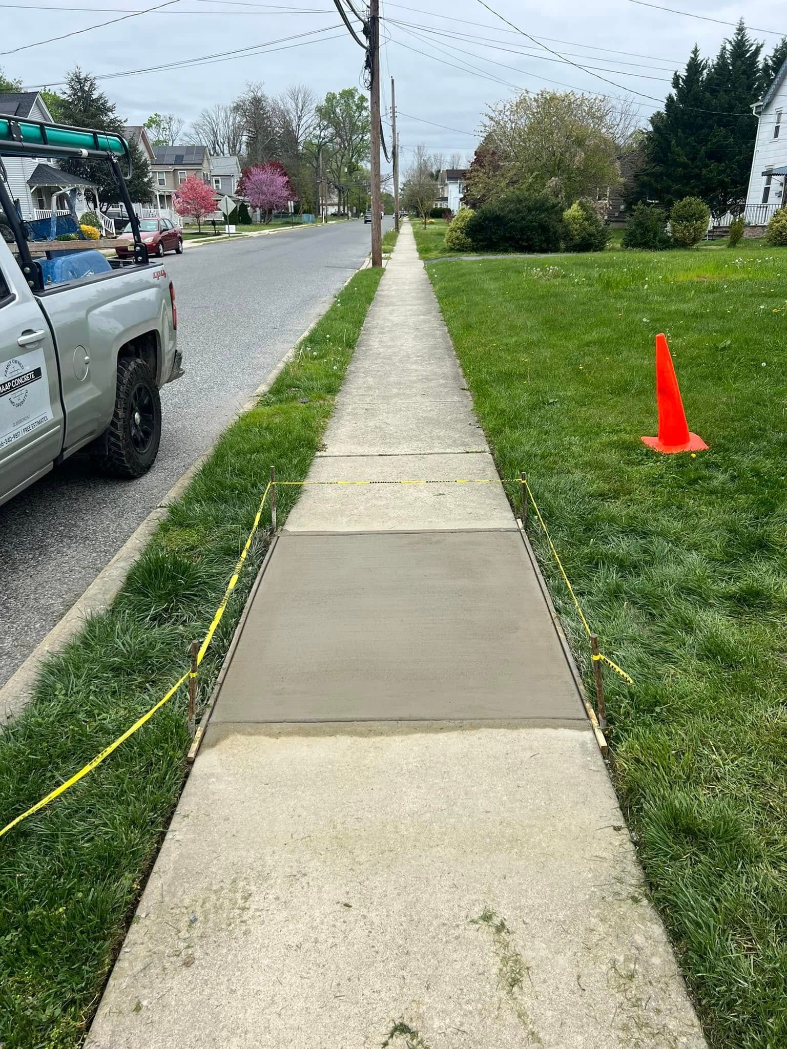 Sidewalk with new concrete section, grass, orange cone, utility truck.