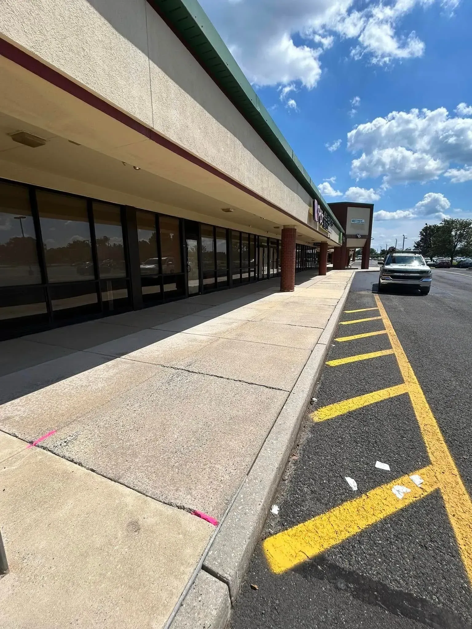 Exterior of a strip mall with a concrete sidewalk, empty storefronts, and yellow parking lines. Sunny day.
