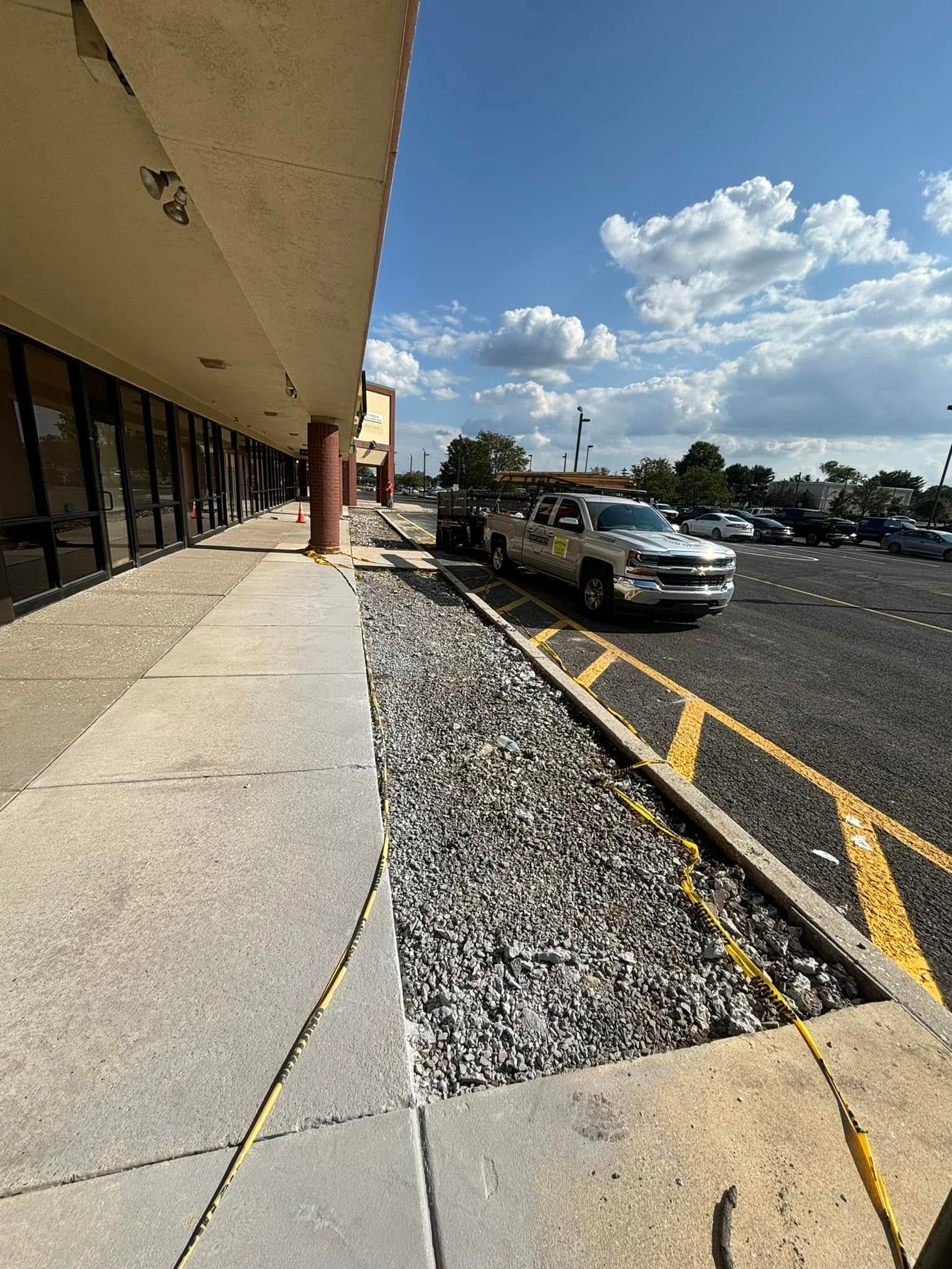 A silver truck parked near a sidewalk and gravel area at a shopping center on a sunny day.
