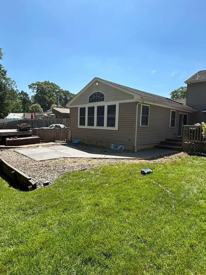 Backyard with a tan-sided house, windows, and a patio with landscaping stones. Green grass and a blue sky.