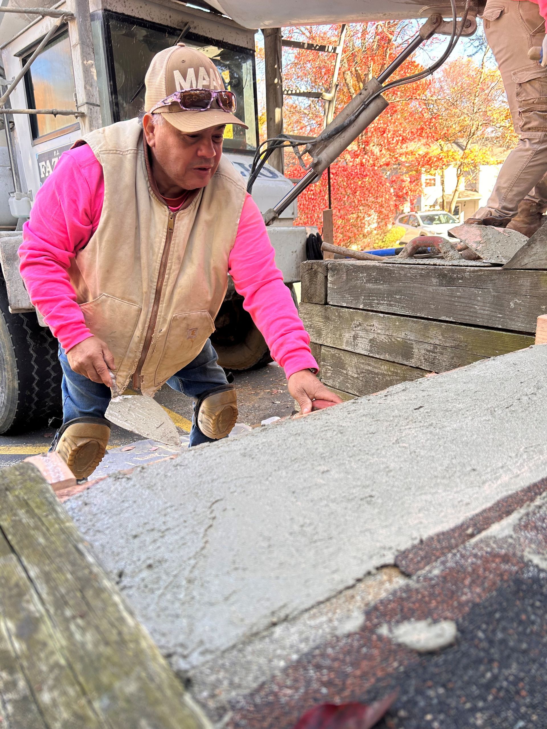 Man in pink sweatshirt, beige vest, and cap spreading cement on a roof with a trowel.