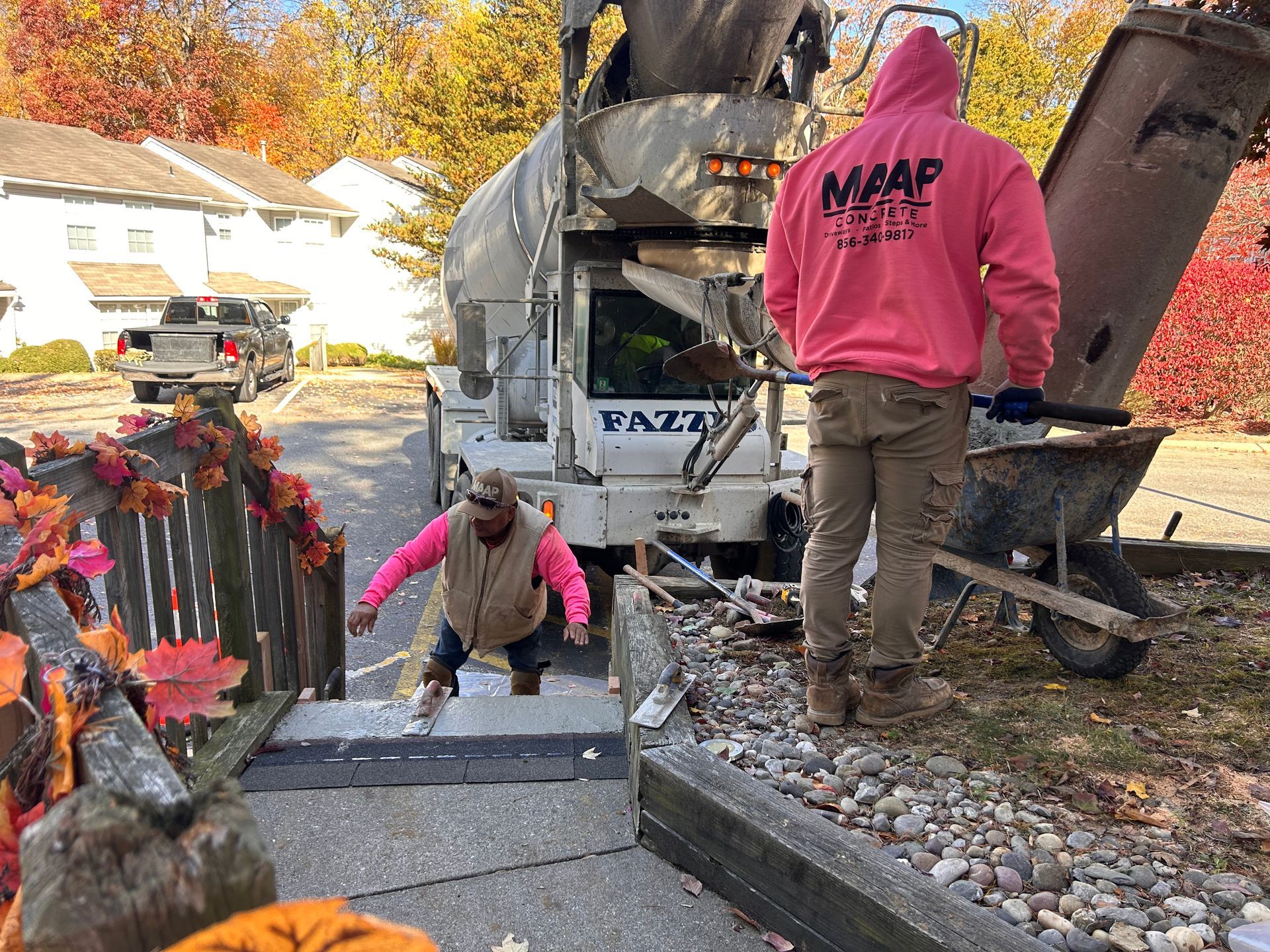 Two construction workers pouring concrete from a truck into a wheelbarrow near a residential area.